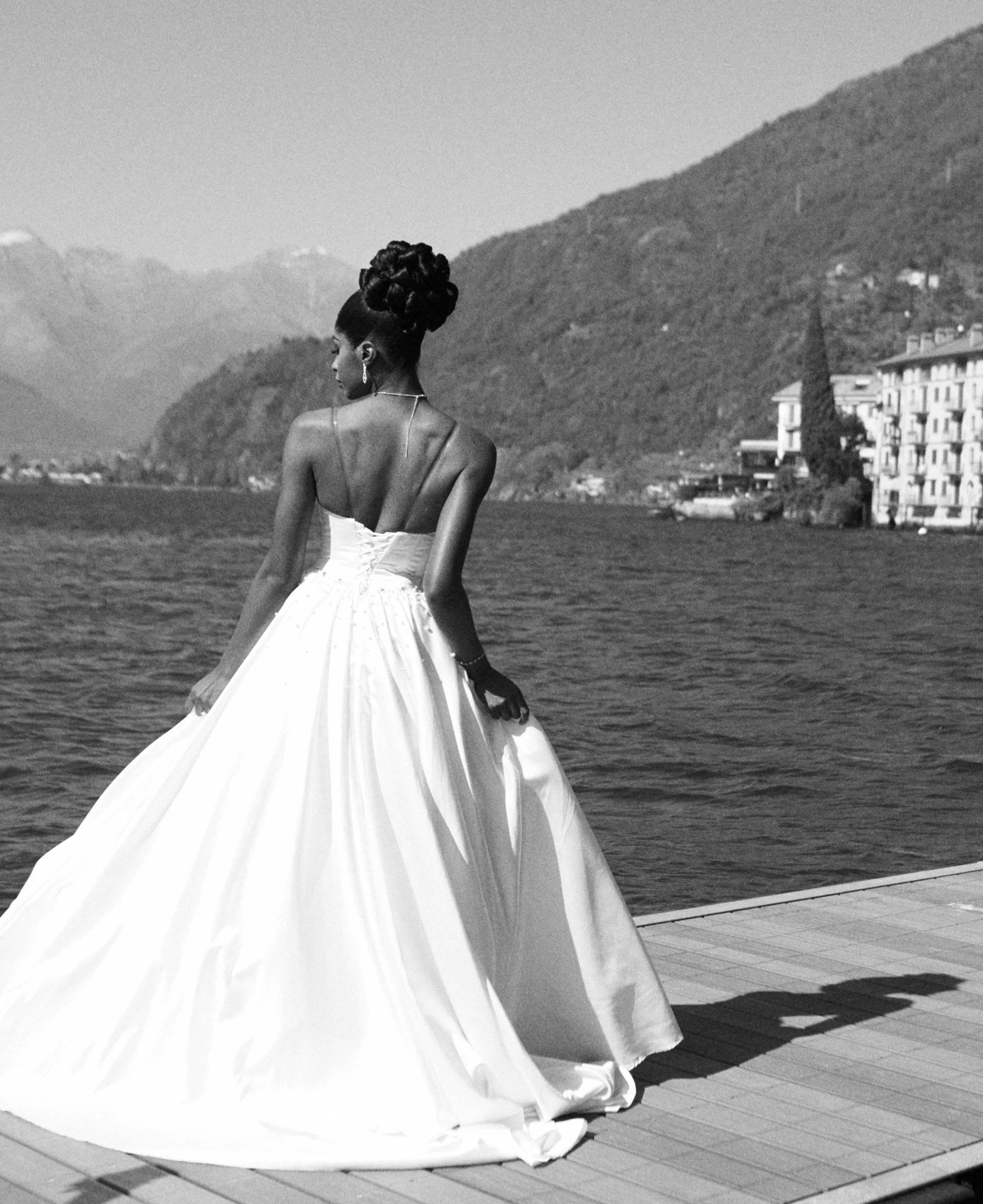 Woman in an elegant strapless wedding dress with a full skirt, standing outdoors by a body of water with mountains in the background, in black and white.