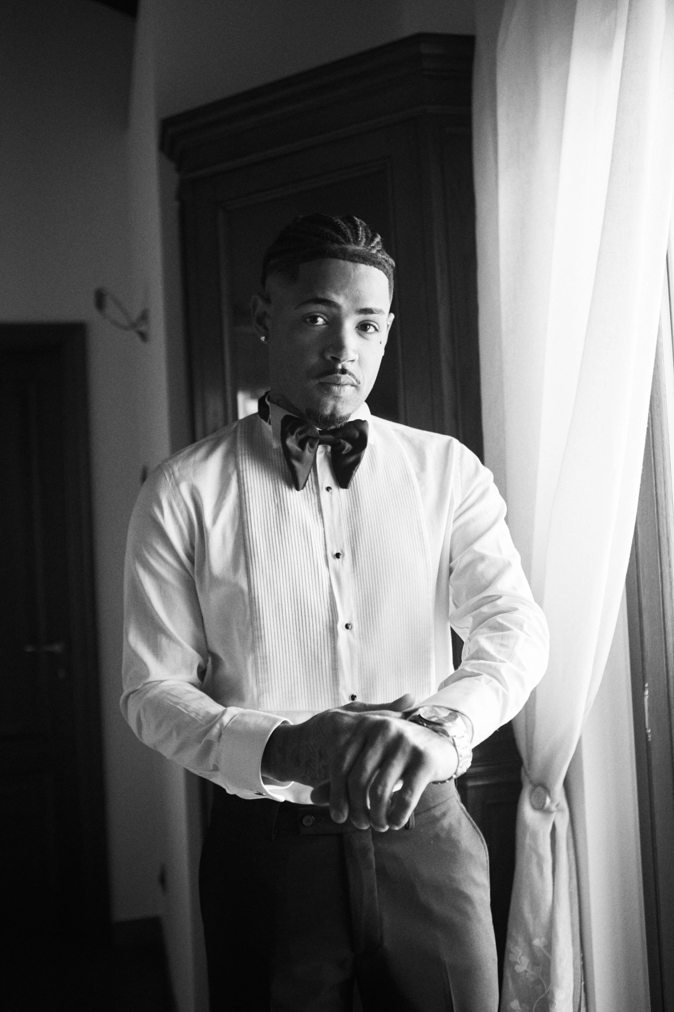 A young man with styled hair, wearing a tuxedo with a bow tie, standing in front of a mirror and taking a selfie in black and white.