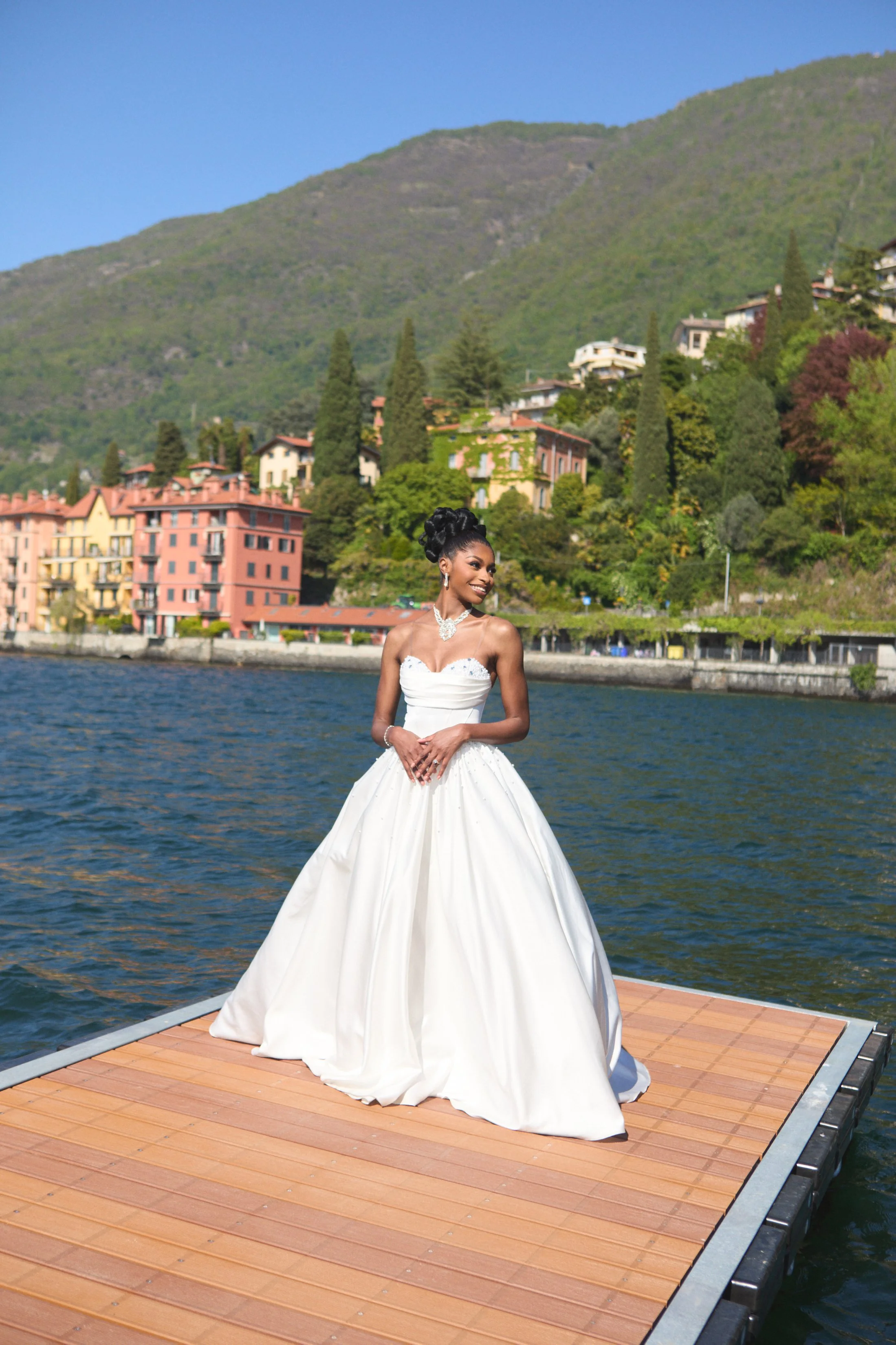 A woman in a white wedding dress is standing on a dock by the water, with colorful houses and green hills in the background.