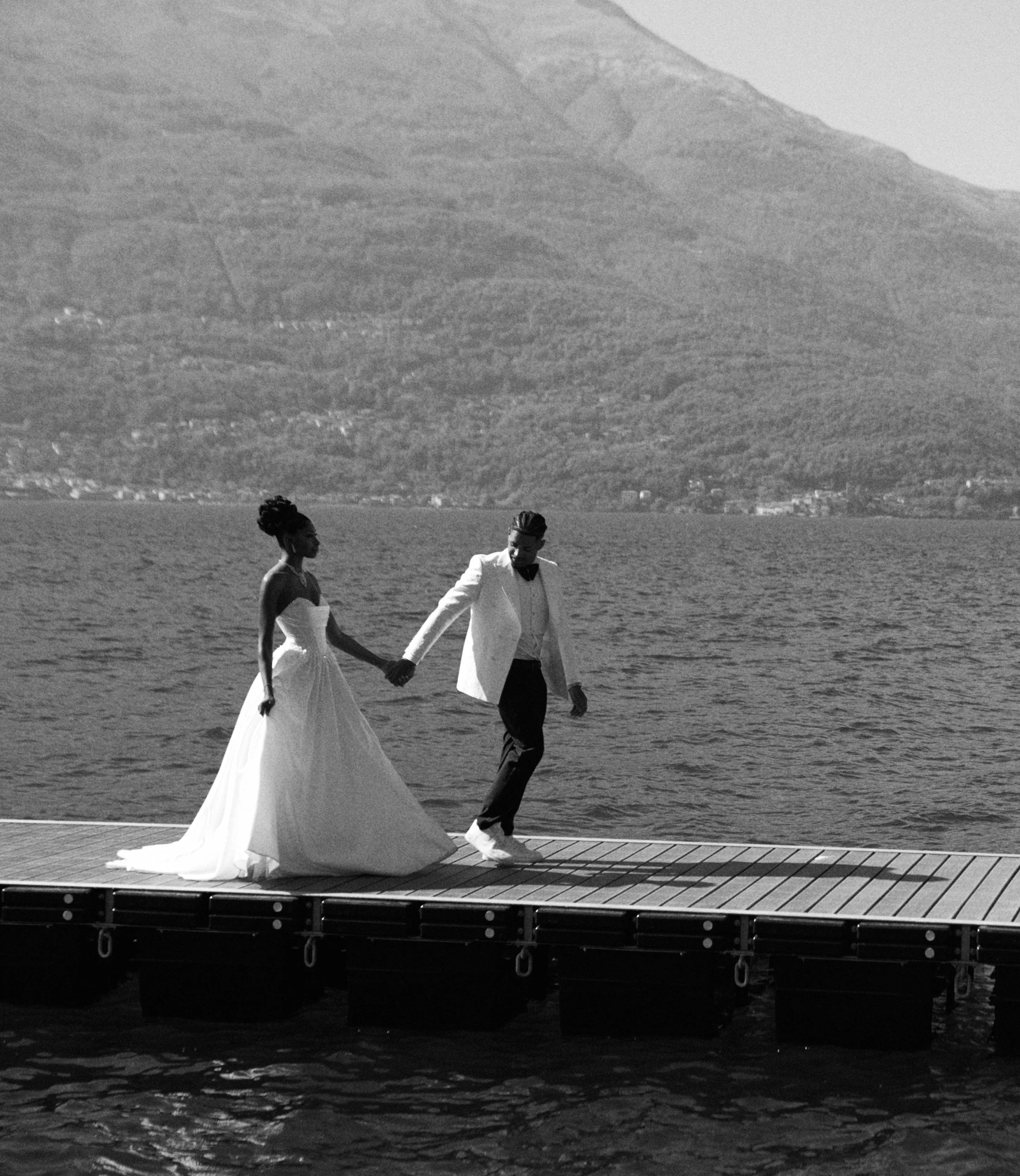 A black and white photo of a couple dressed in formal attire, walking hand in hand on a wooden dock by a body of water with a mountain in the background. The woman is wearing a wedding gown and the man is in a tuxedo.