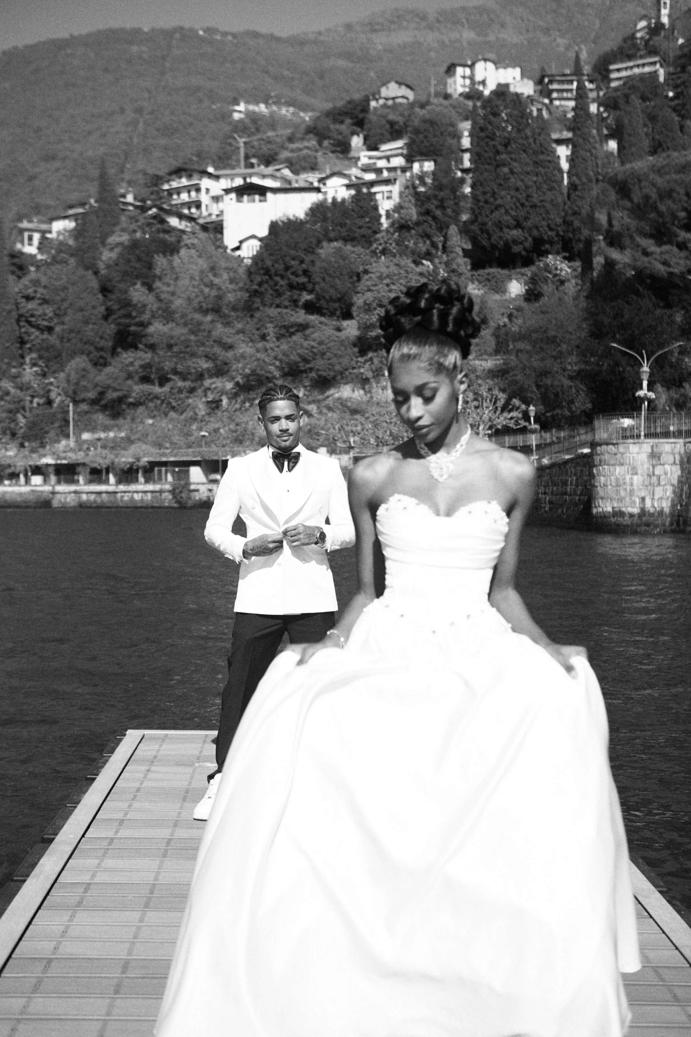 Black and white photo of a couple in wedding attire on a dock by the water, with a hilly residential area in the background.