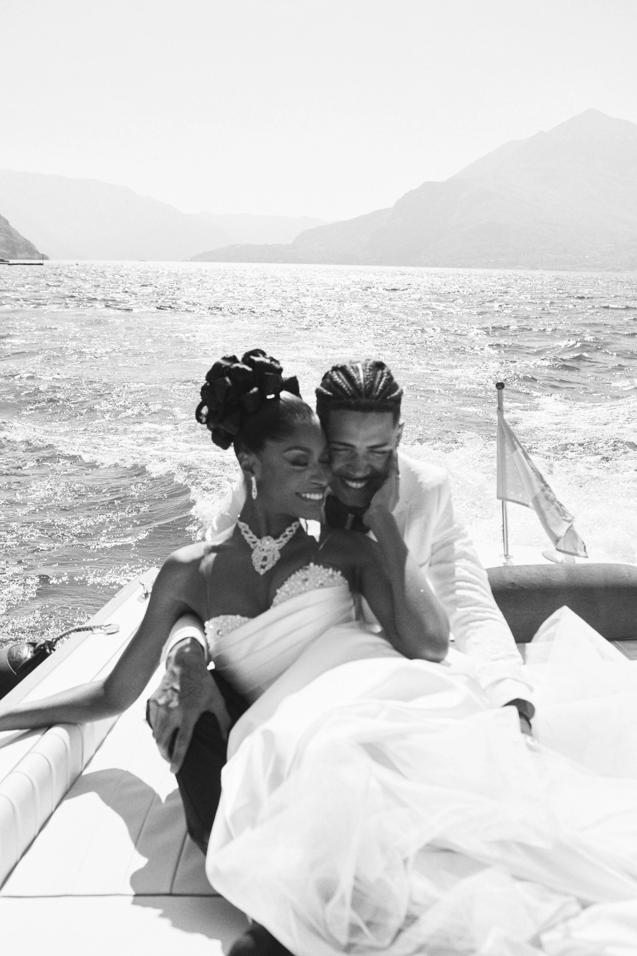 A black and white photo of a smiling couple on a boat, with a scenic lake and mountains in the background, during daytime.