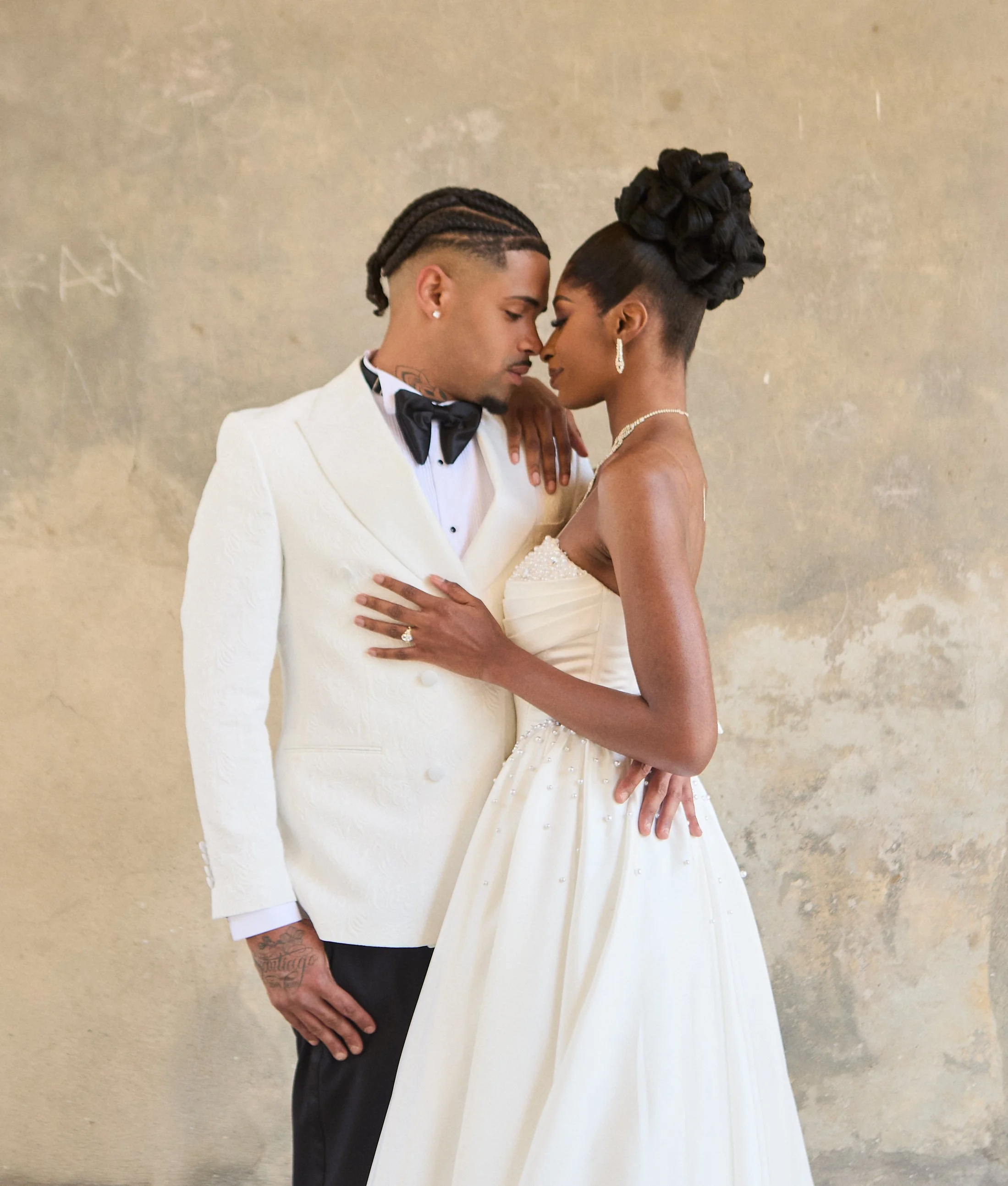 A couple with dark skin, dressed in formal wedding attire, standing close with their foreheads together against a plain wall. The man wears a white tuxedo with a black bow tie, and has braided hair and tattoos on his left hand. The woman has an elabo