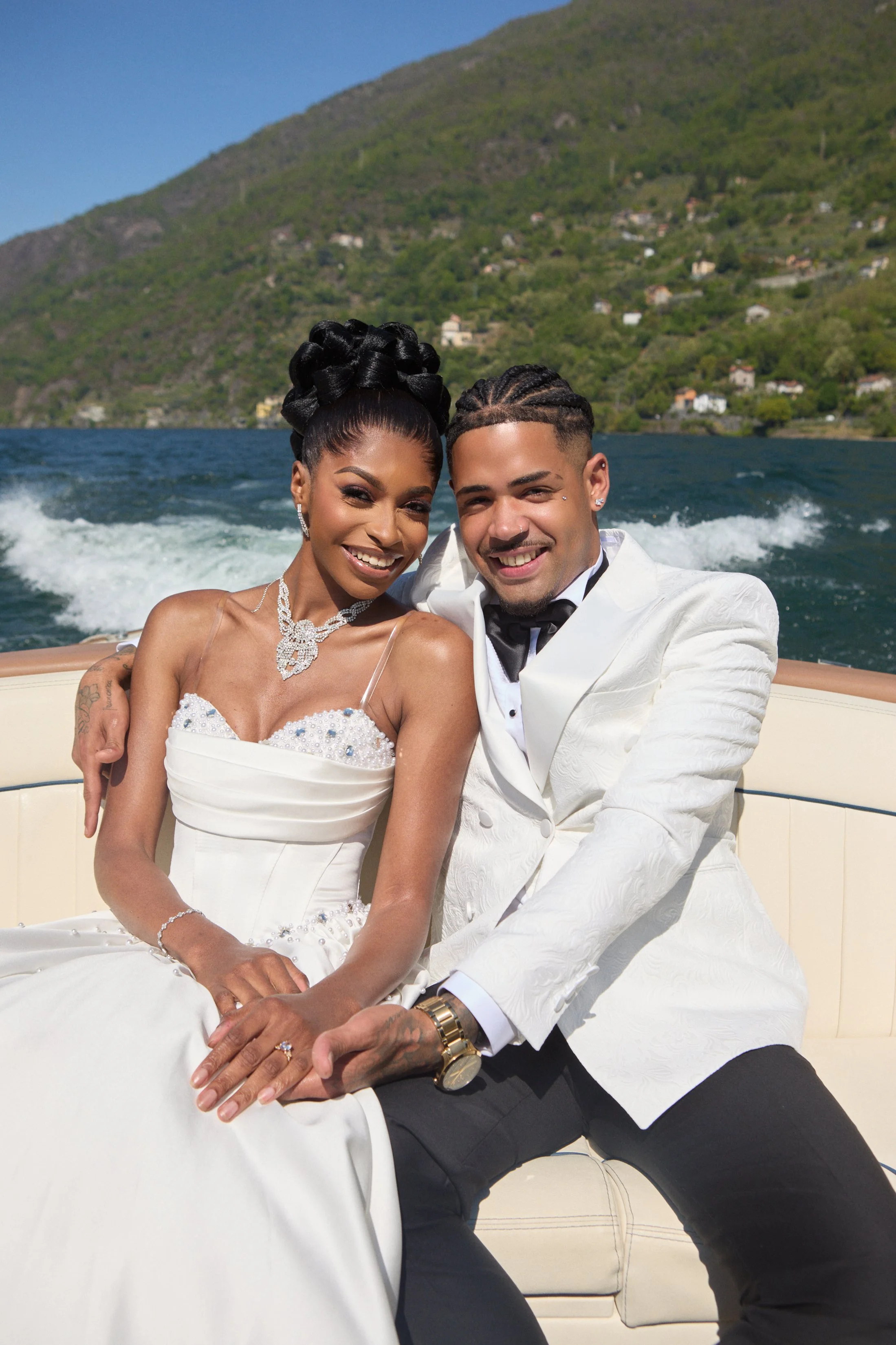 A happy couple dressed in wedding attire sitting on a boat with a scenic hillside and lake in the background.