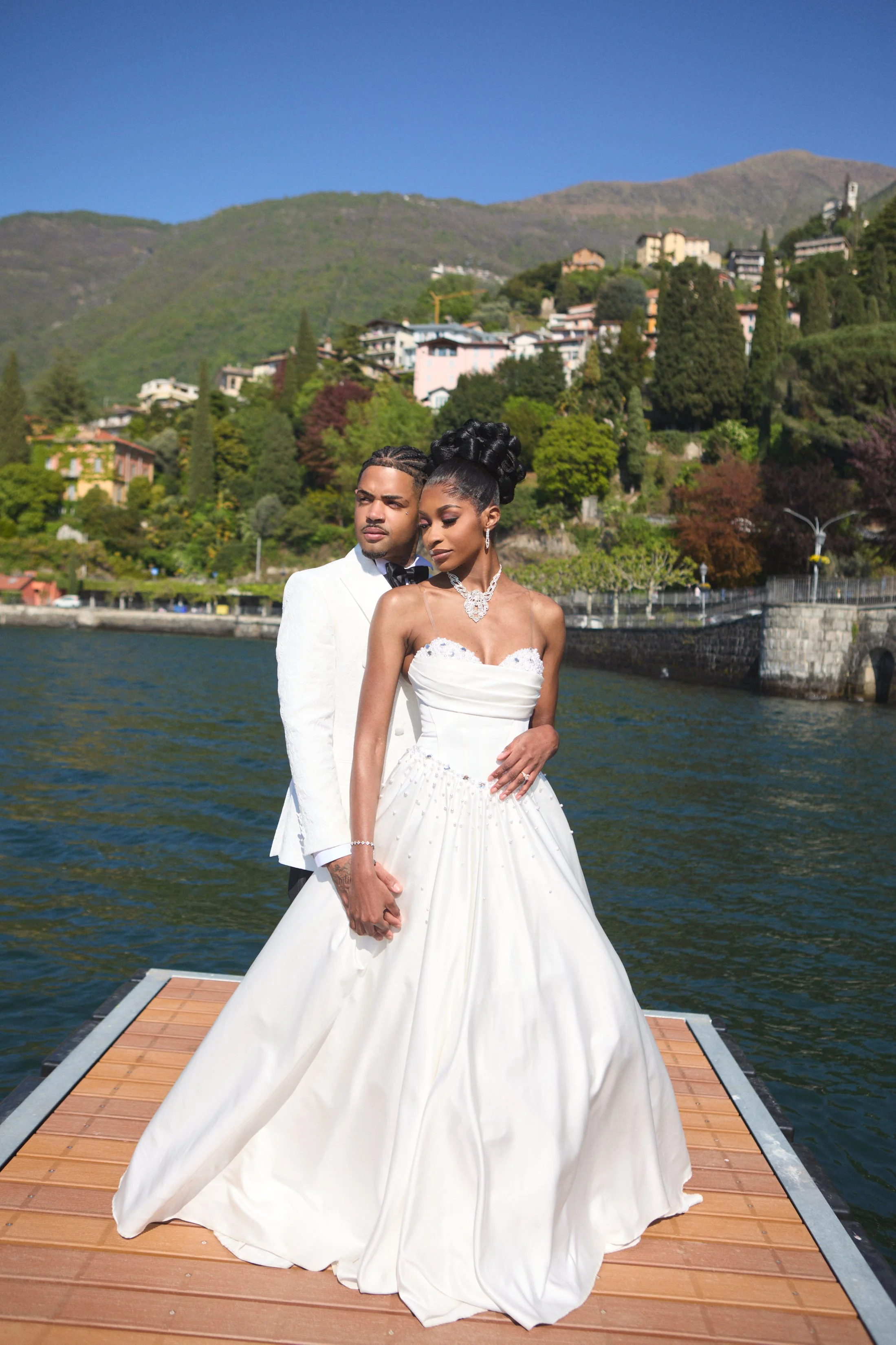 A couple dressed in wedding attire standing on a dock by the water with a hilly, residential area and blue sky in the background.
