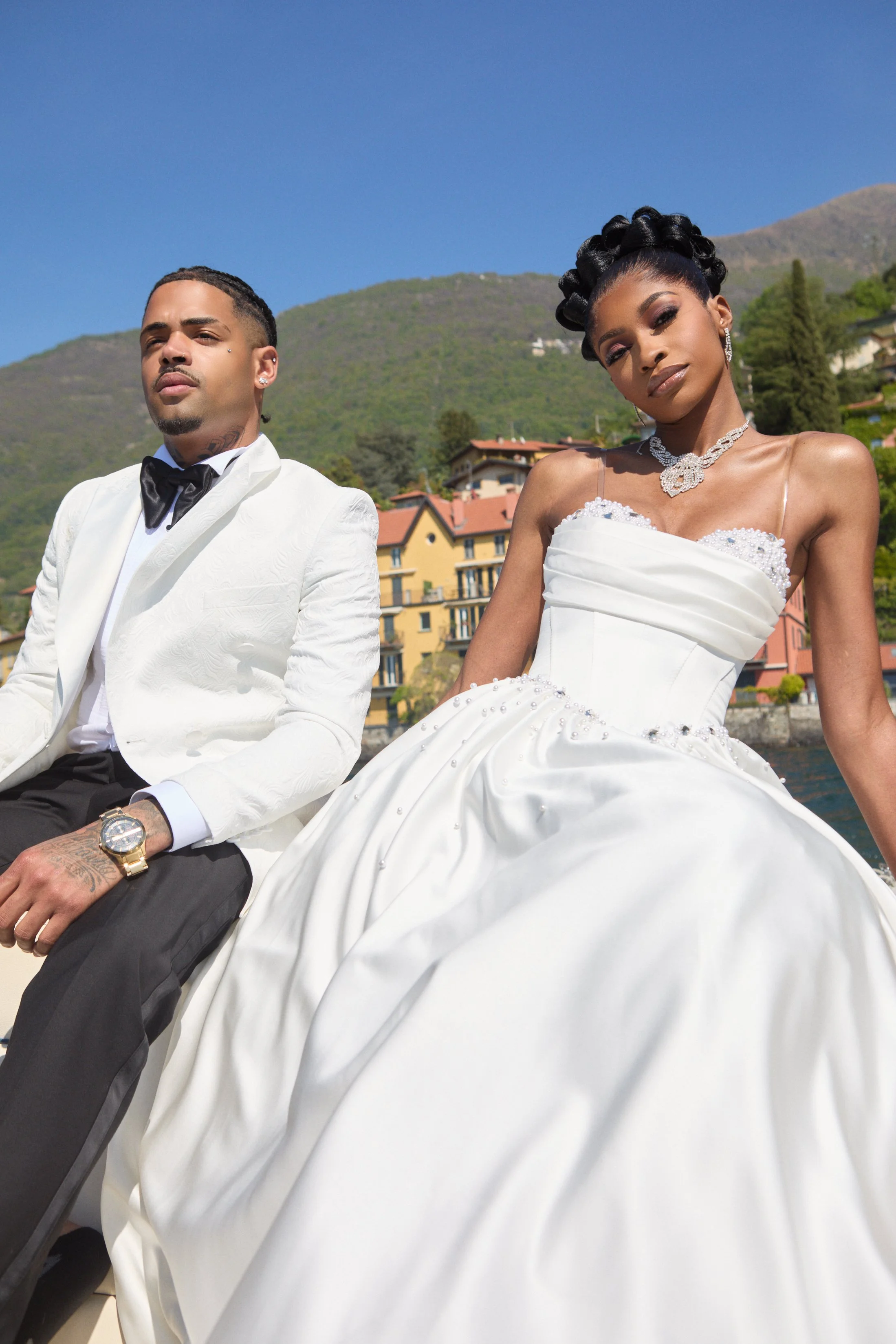 A man and woman dressed in formal wedding attire sitting outdoors with a hillside and buildings in the background. The man is in a white tuxedo with a black bow tie, and the woman is in a white wedding gown with pearl embellishments and jewelry.