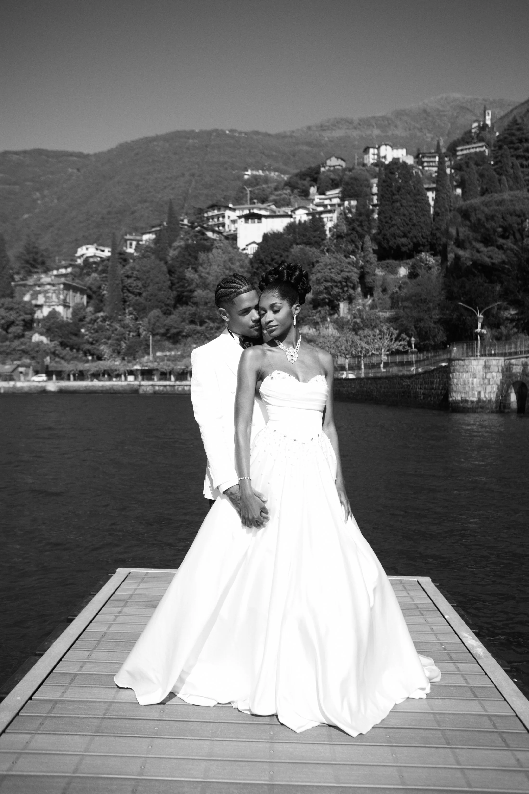A couple dressed in wedding attire standing on a dock with a lake and hillside houses in the background, in a black and white photograph.