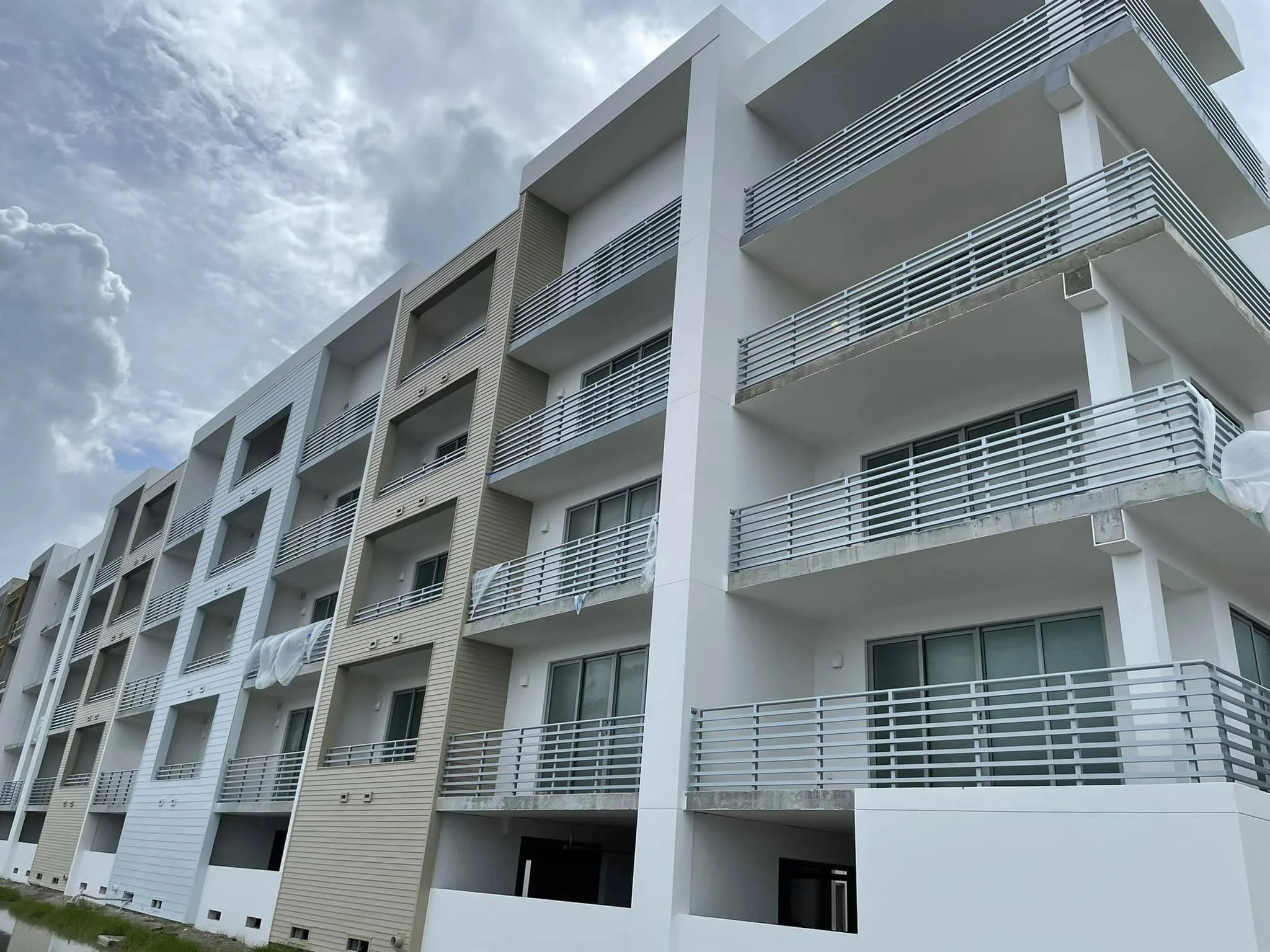 Modern white apartment building with multiple balconies, metal railings, and large windows under a partly cloudy sky.