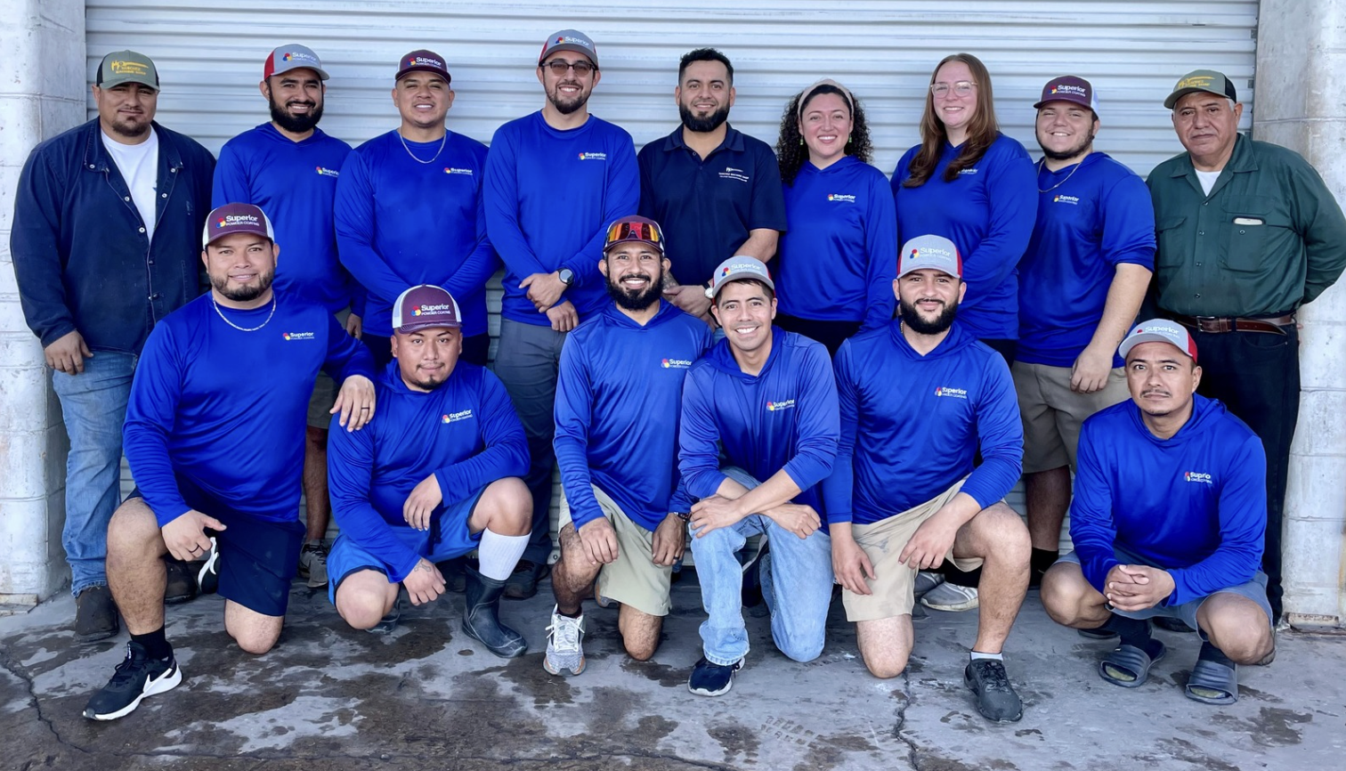 Group of people posing for a photo in front of a metal rolling door, some wearing matching blue shirts with a logo, some wearing caps, standing outdoors.