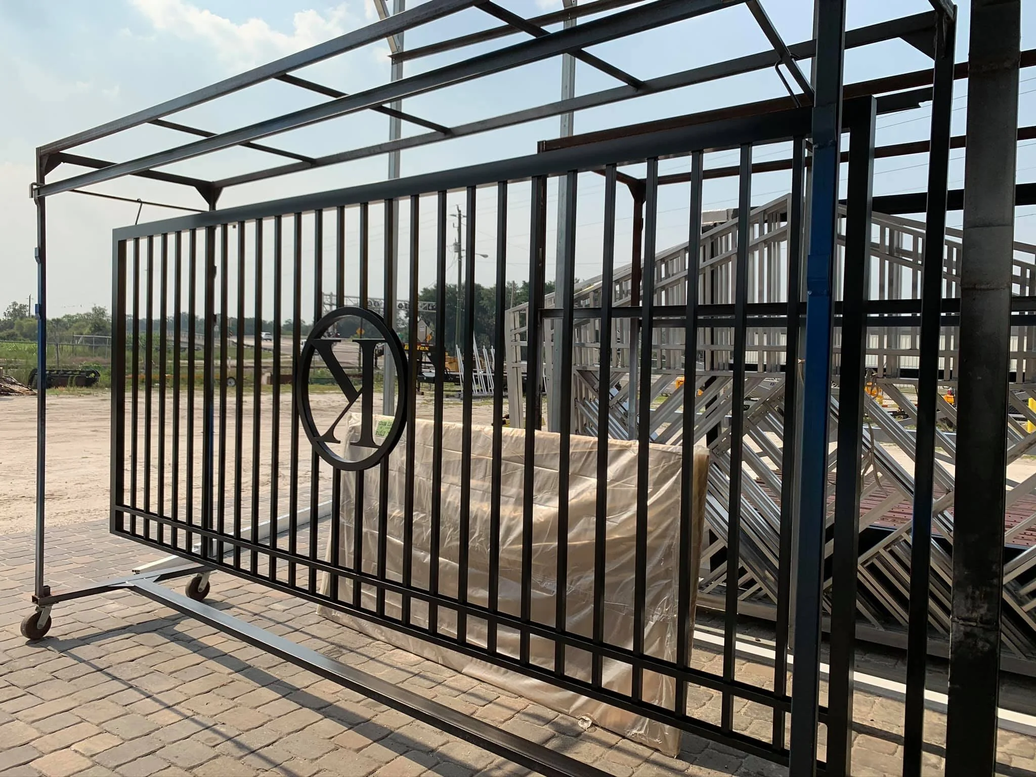 A black metal gate with a circular logo in the center and wheels at the bottom, standing outdoors on a paved surface with construction materials and metal stairs in the background.