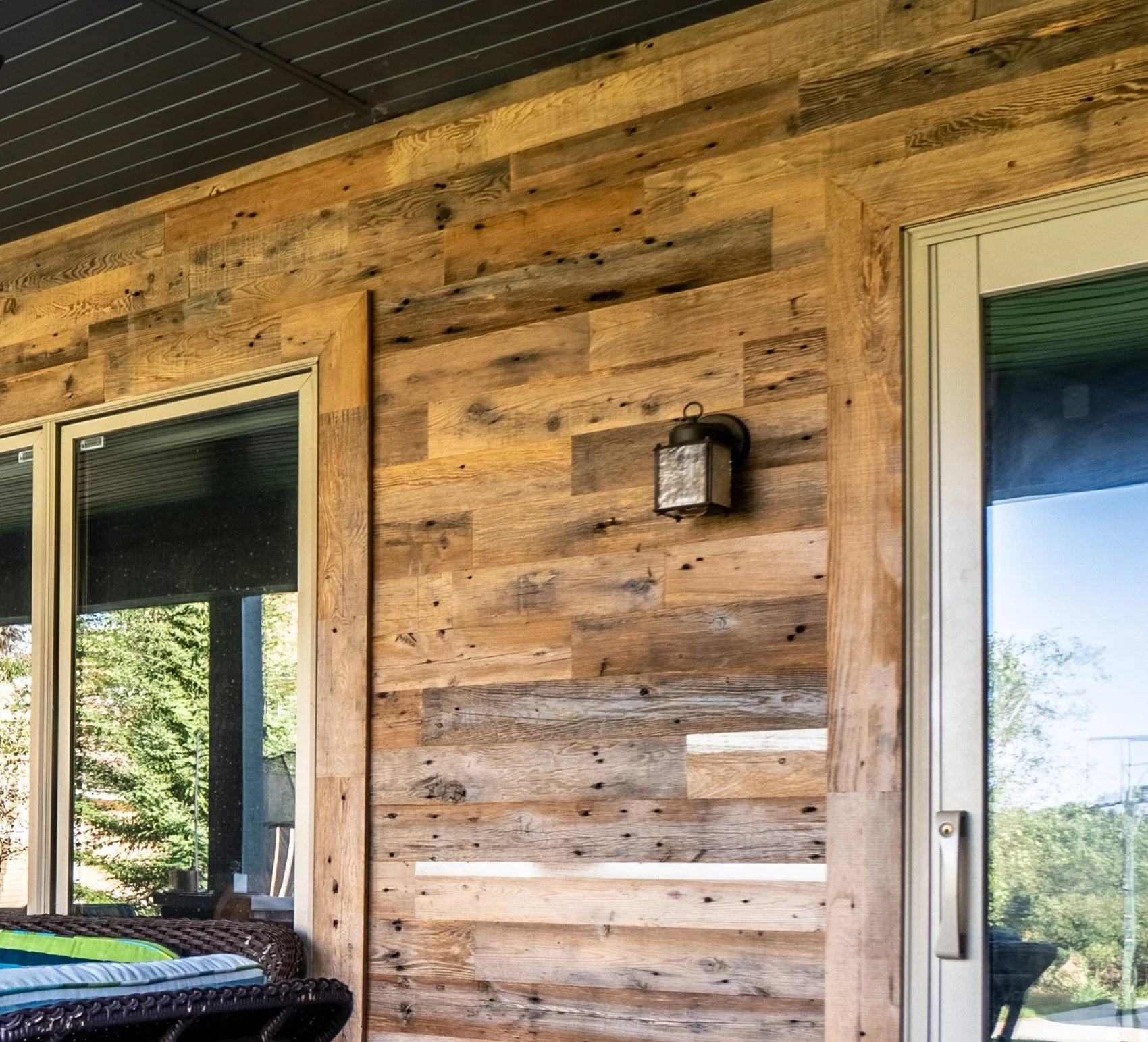 Wood-paneled exterior wall with a black outdoor wall light fixture, adjacent to sliding glass doors, and a reflection of greenery in the glass.