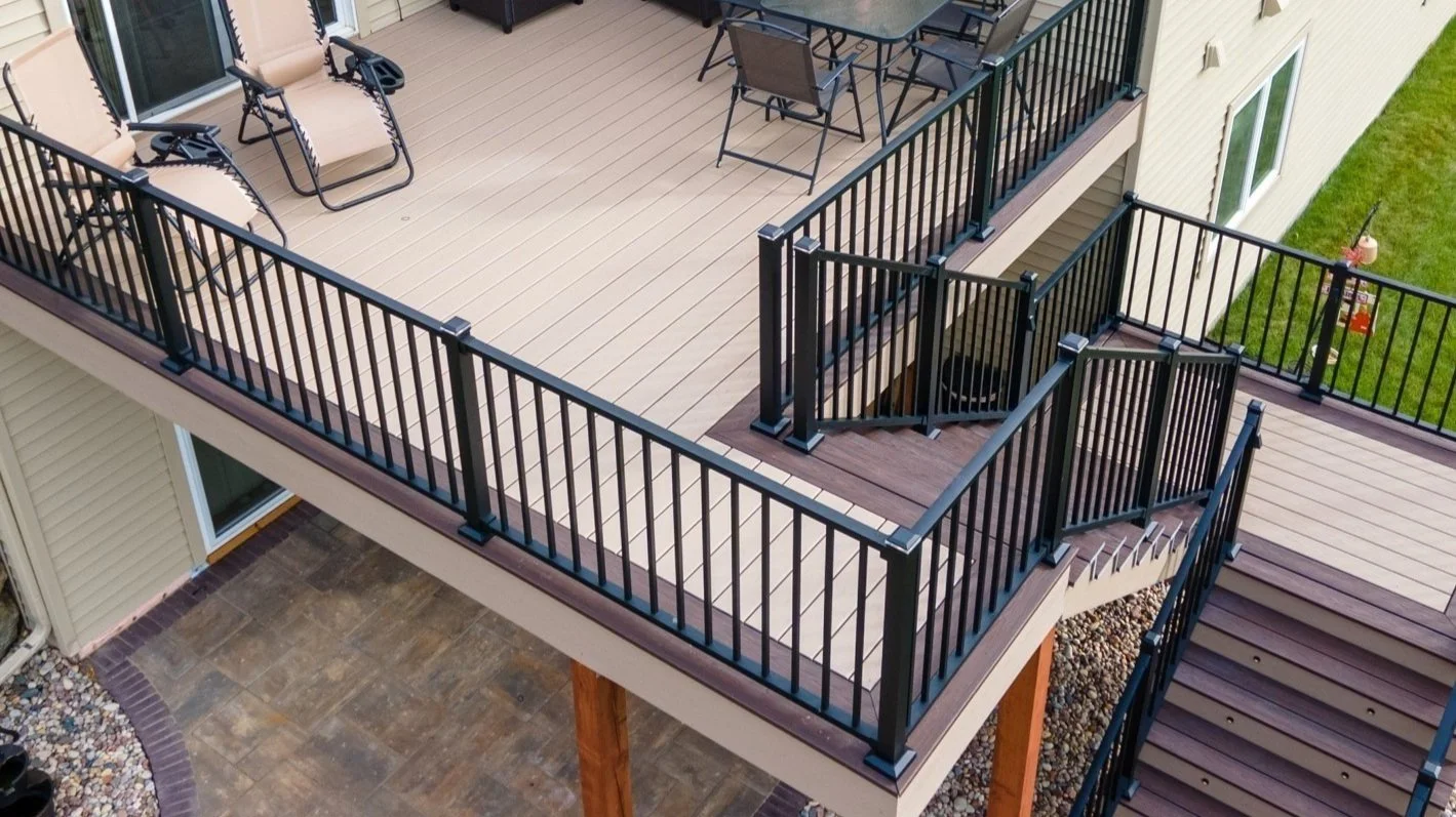 An outdoor balcony with beige decking, black metal railing, outdoor furniture, and stairs leading down to a yard.