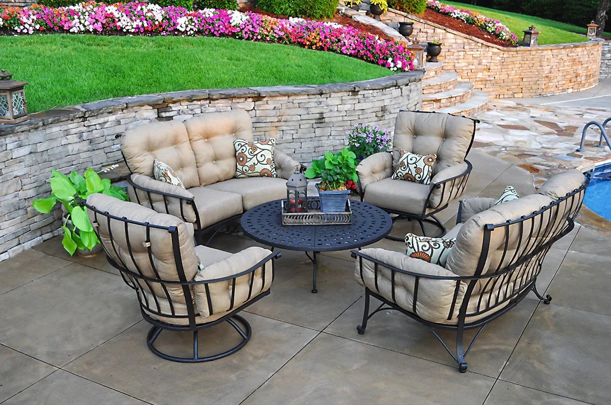 five piece wrought iron lounge set with tan cushions around round coffee table on hardscape next to stone wall and garden