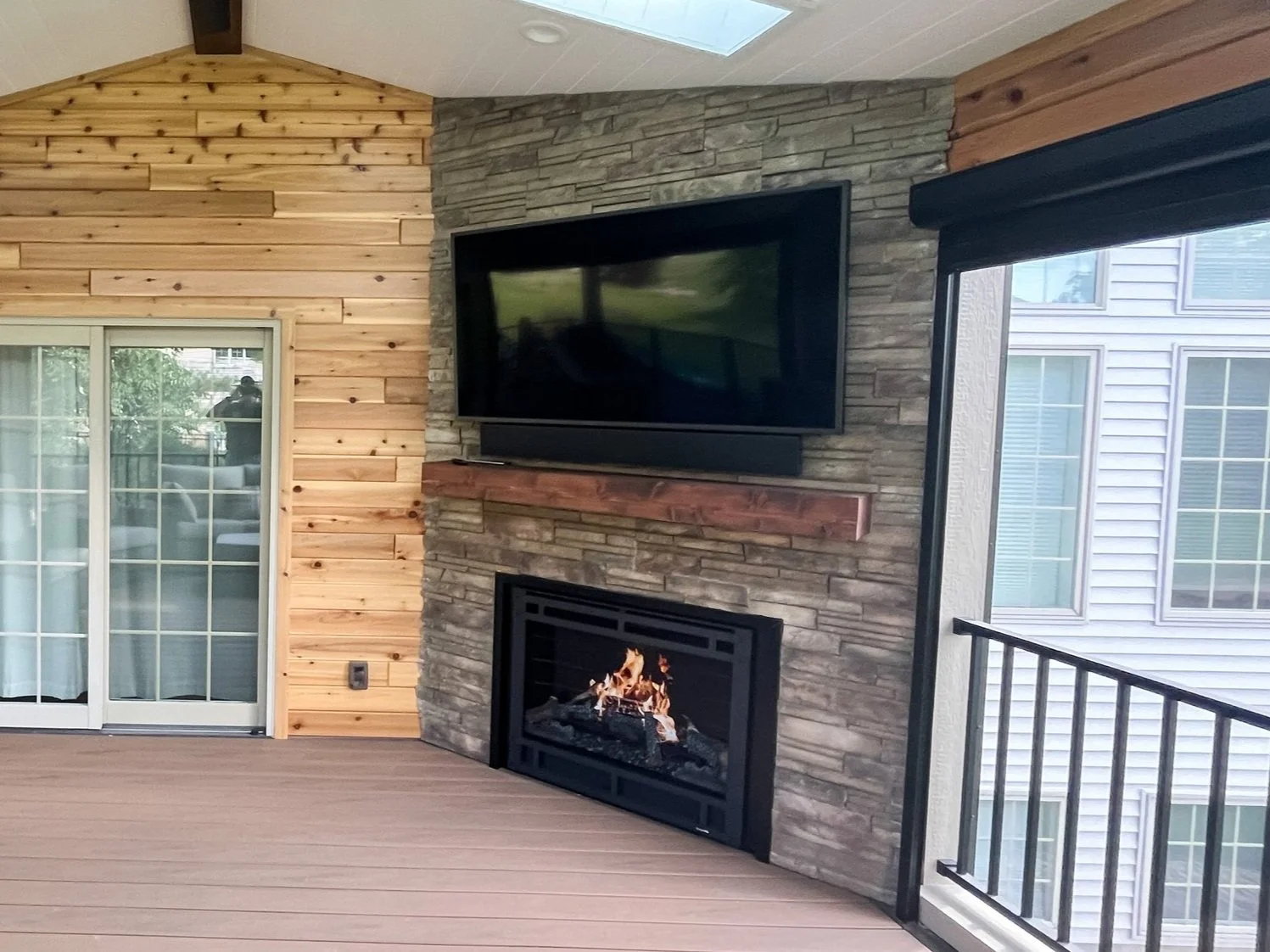 Outdoor patio area with sliding glass door, wooden wall, stone fireplace, mounted flat-screen TV, and railing.