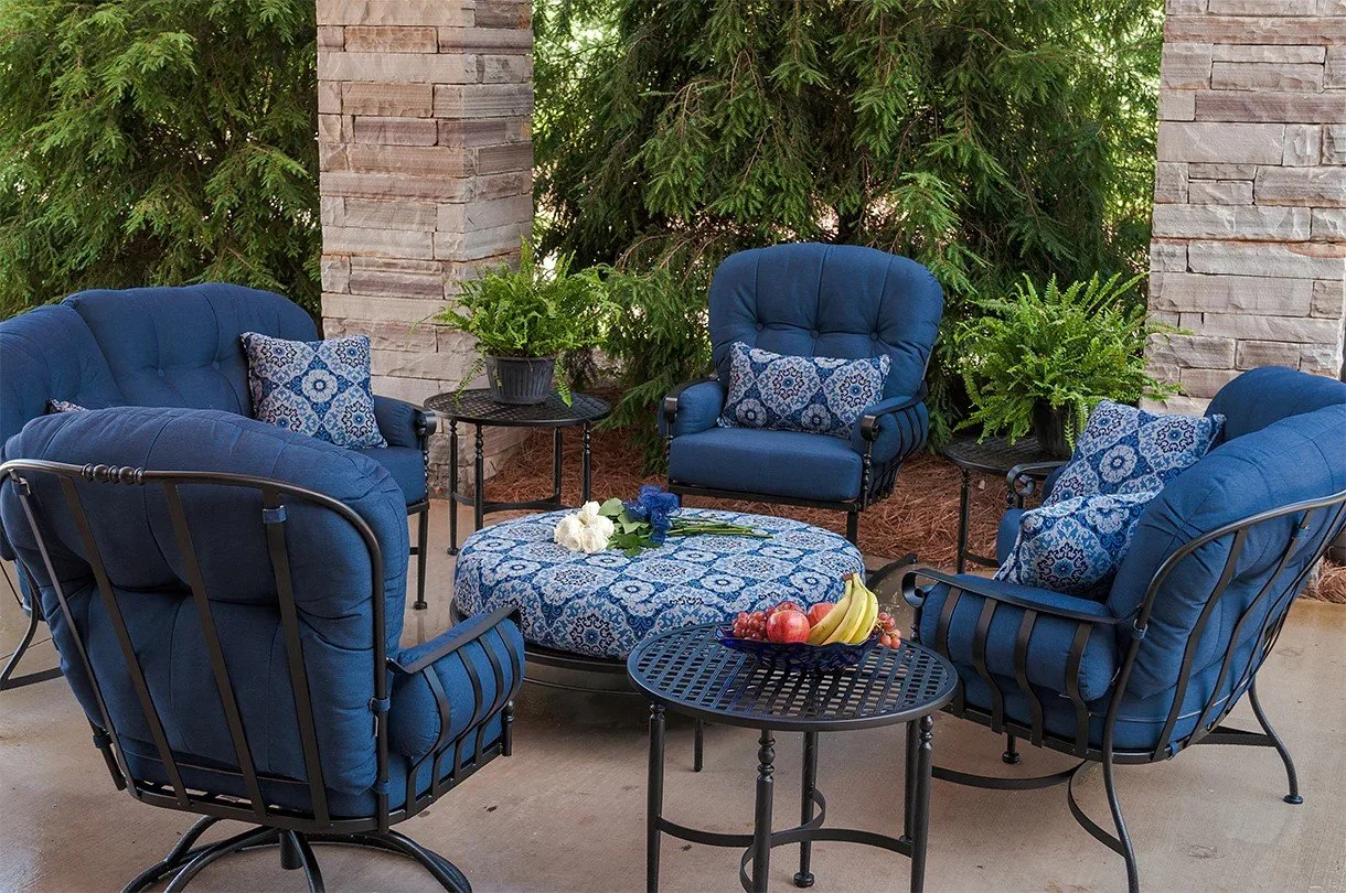 wrought iron deep seated lounge chairs with blue cushions around patterned blue ottoman next to end tables on concrete surface