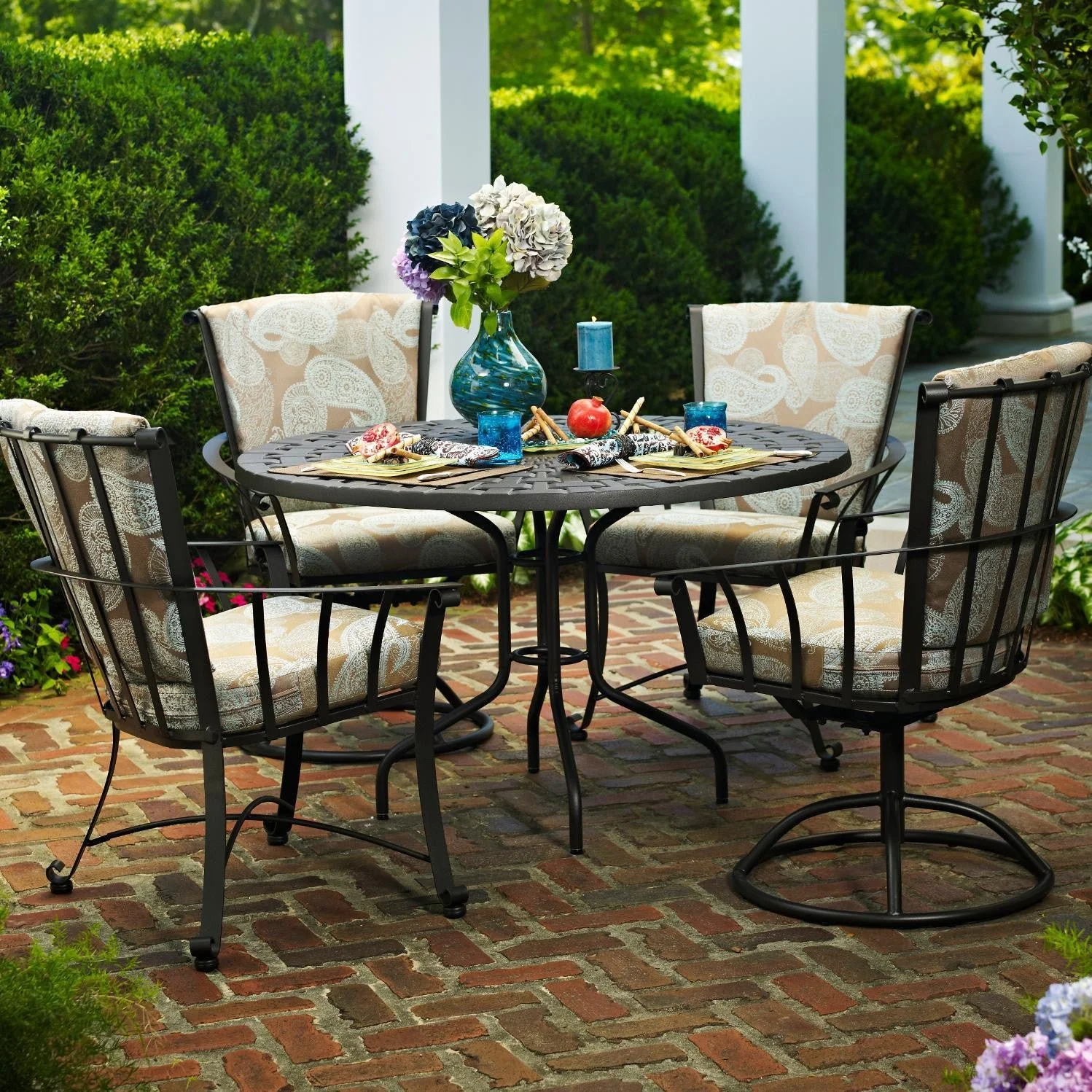 four wrought iron chairs with brown and white patterned cushions around round table on brick surface in front of bushes