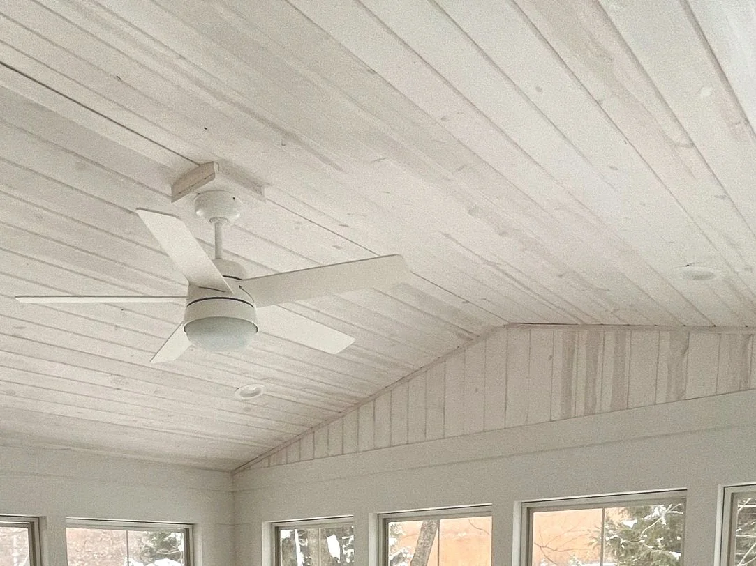 White ceiling with a modern ceiling fan and large windows showing a view of trees outside.