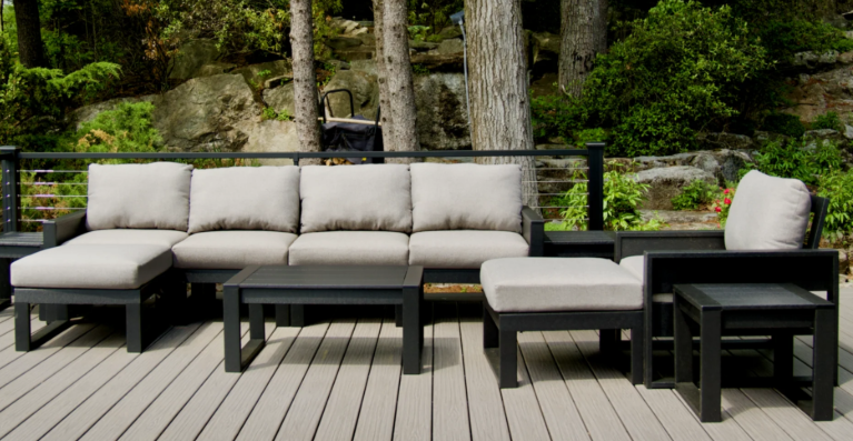 Outdoor patio furniture set with a large beige cushioned sofa, two matching cushioned chairs, and a black coffee table, set on a wooden deck with trees and rocks in the background.