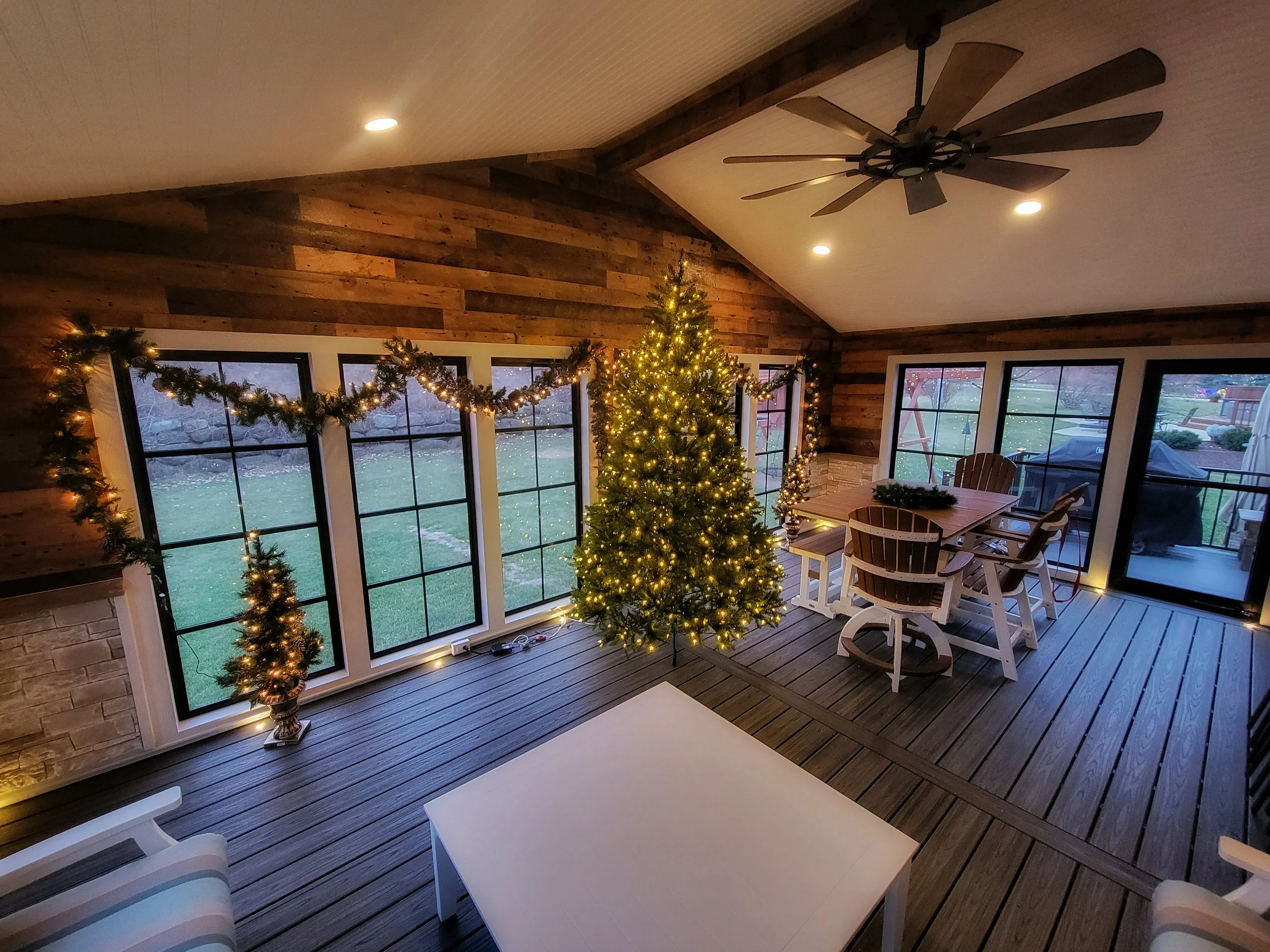A decorated living room with a Christmas tree, string lights, and garlands, overlooking a backyard through large windows. There is a wooden ceiling fan and a dining table with chairs near the windows.