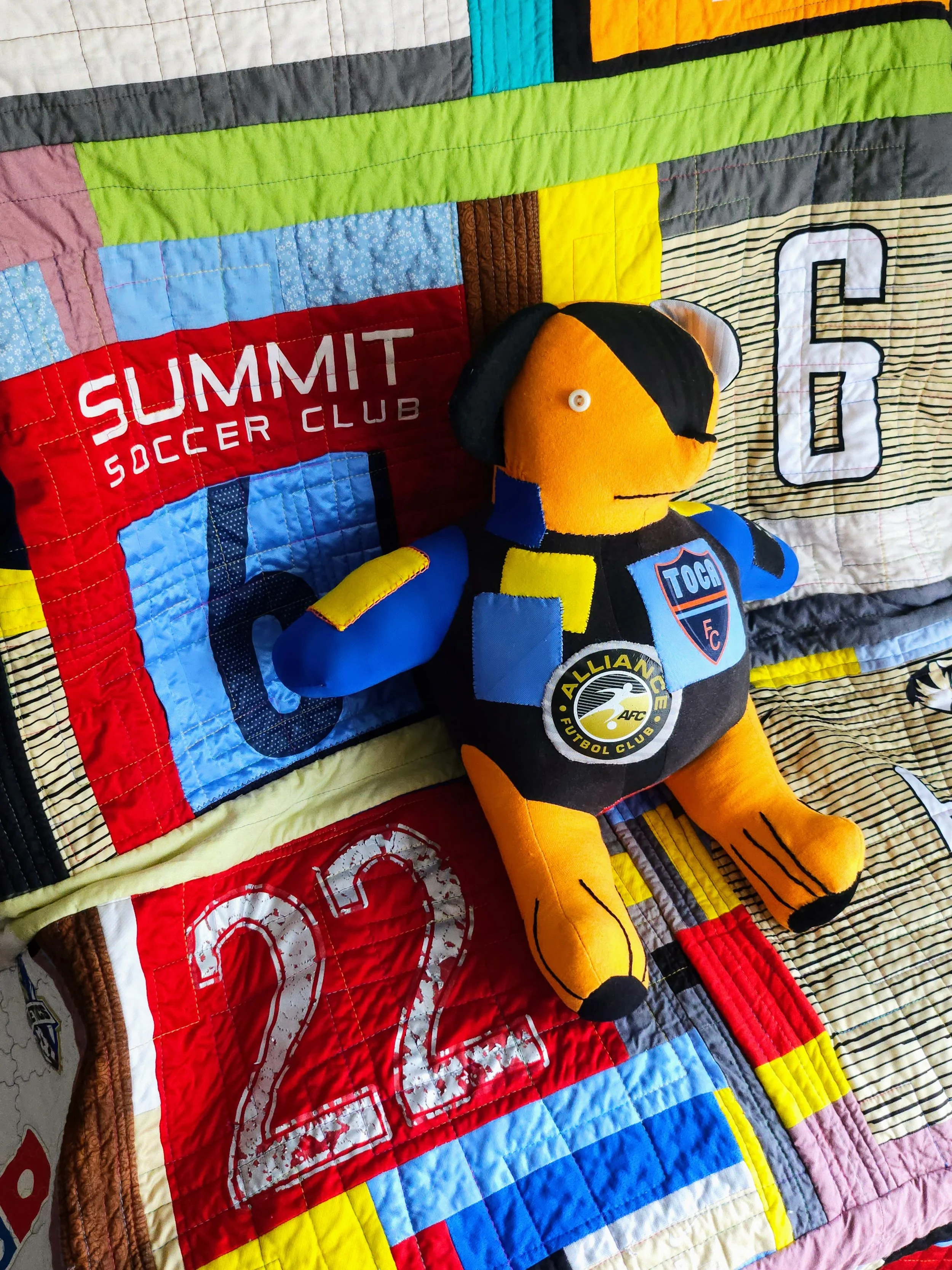 A patchwork stuffed bear sitting on a patchwork quilt.