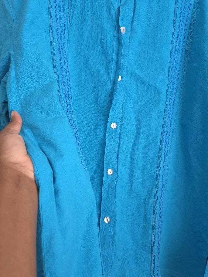 A hand touching a hanging men's blue collar shirt with buttons, pleats and embroidery.