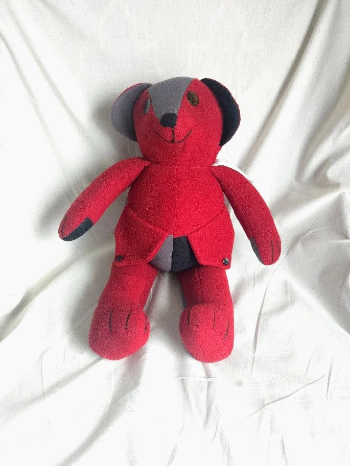 A red and black upcycled stuffed teddy bear with pockets in the front, sitting on a neutral background.