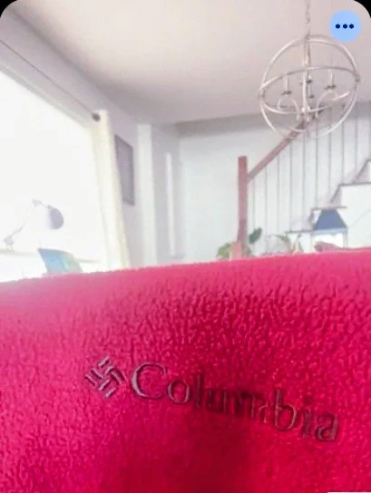Screenshot of extreme close-up of textured red fleece fabric featuring a black embroidered Columbia logo. The background shows a softly blurred room with a staircase.