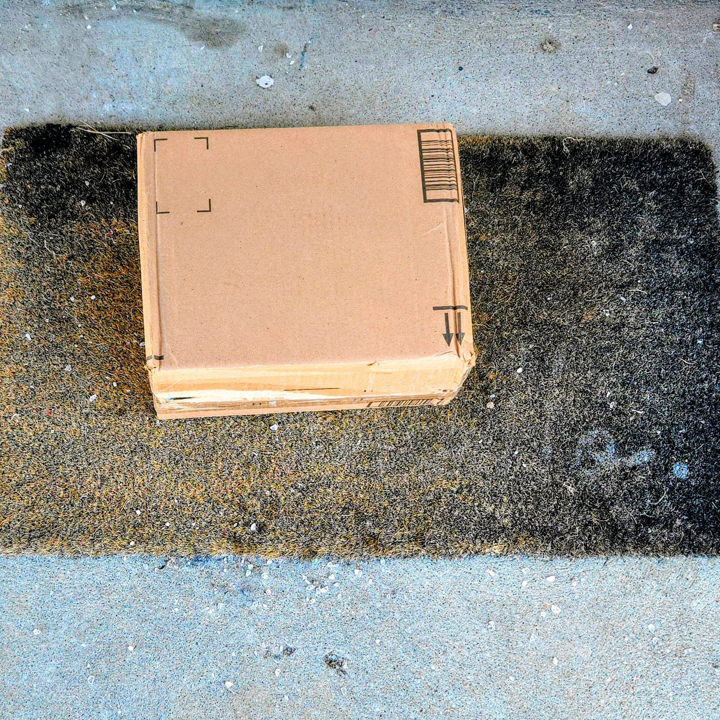 A small, rectangular brown cardboard package wrapped tightly in brown packing tape. It is placed on a textured, grey indoor surface with an out-of-focus background.