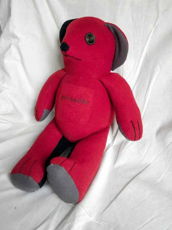 A handmade red and black fleece stuffed teddy bear with the Columbia logo embroidered in the center, sitting on a neutral background.