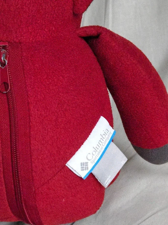 A zipper stitched in the back of a red fleece stuffed teddy bear, with the Columbia clothing tag on the bottom right of the stuffed bear.