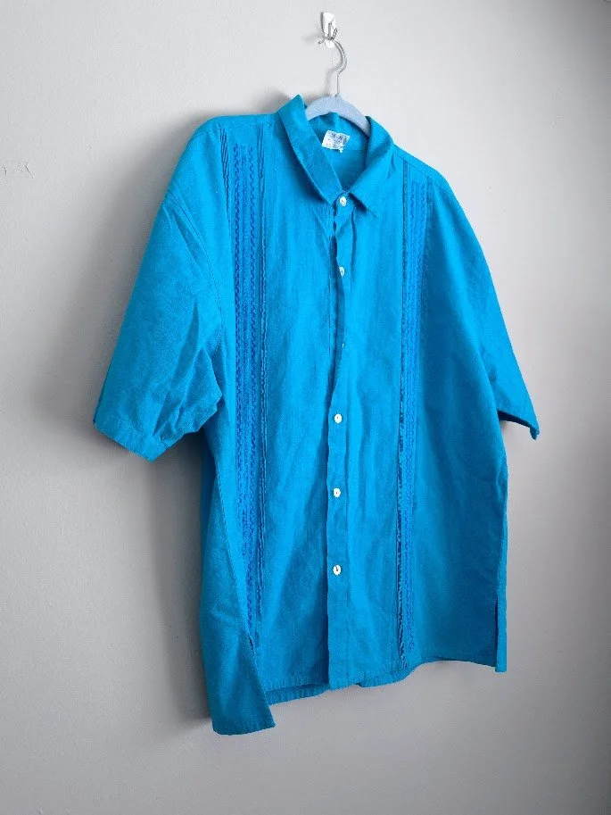 A men's blue embroidery and pleated shirt hanging on a neutral wall.