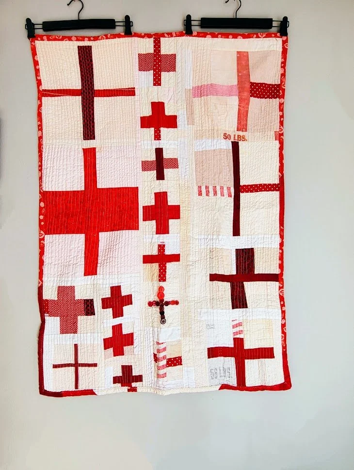 A red and white quilt with crosses hanging on a white wall.