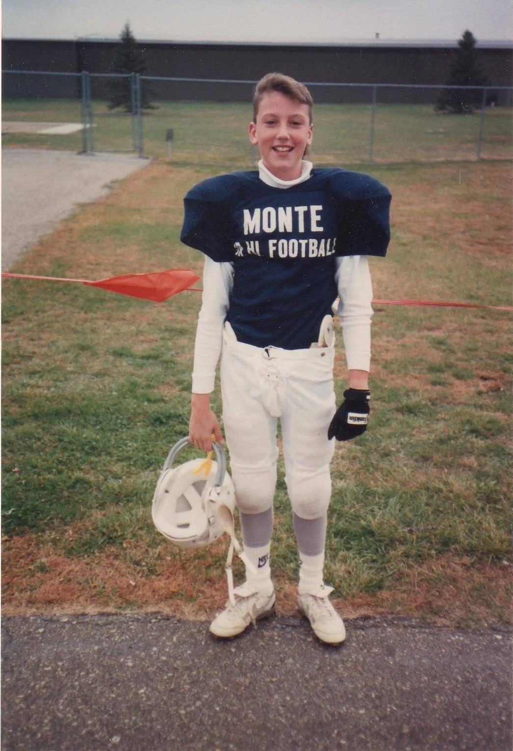 A young Jerrid Sebesta in his American football player in uniform holding a helmet, standing on a field with a fence and building in the background.