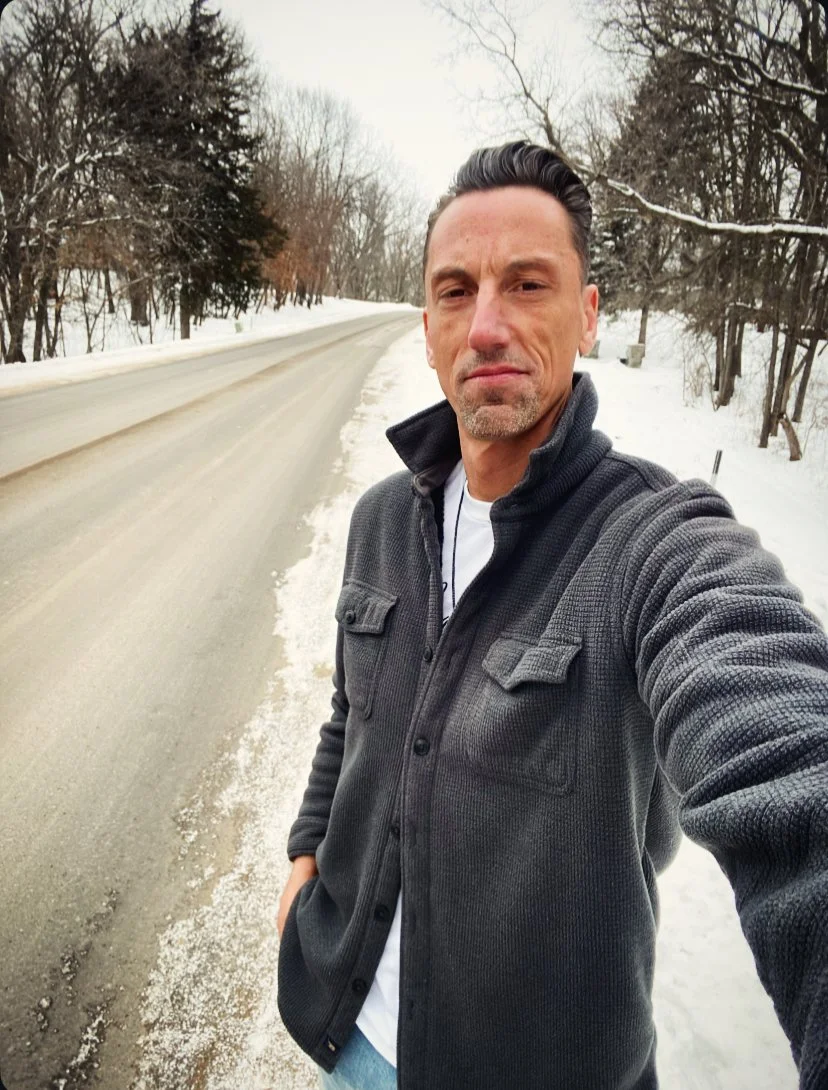 Jerrid Sebesta taking a selfie outdoors on a snowy road during daytime, with trees on both sides.
