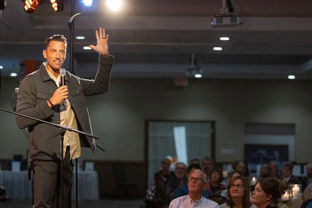 Jerrid Sebesta is speaking on stage holding a microphone, waving to the audience, in a dimly lit room with seated attendees watching him.