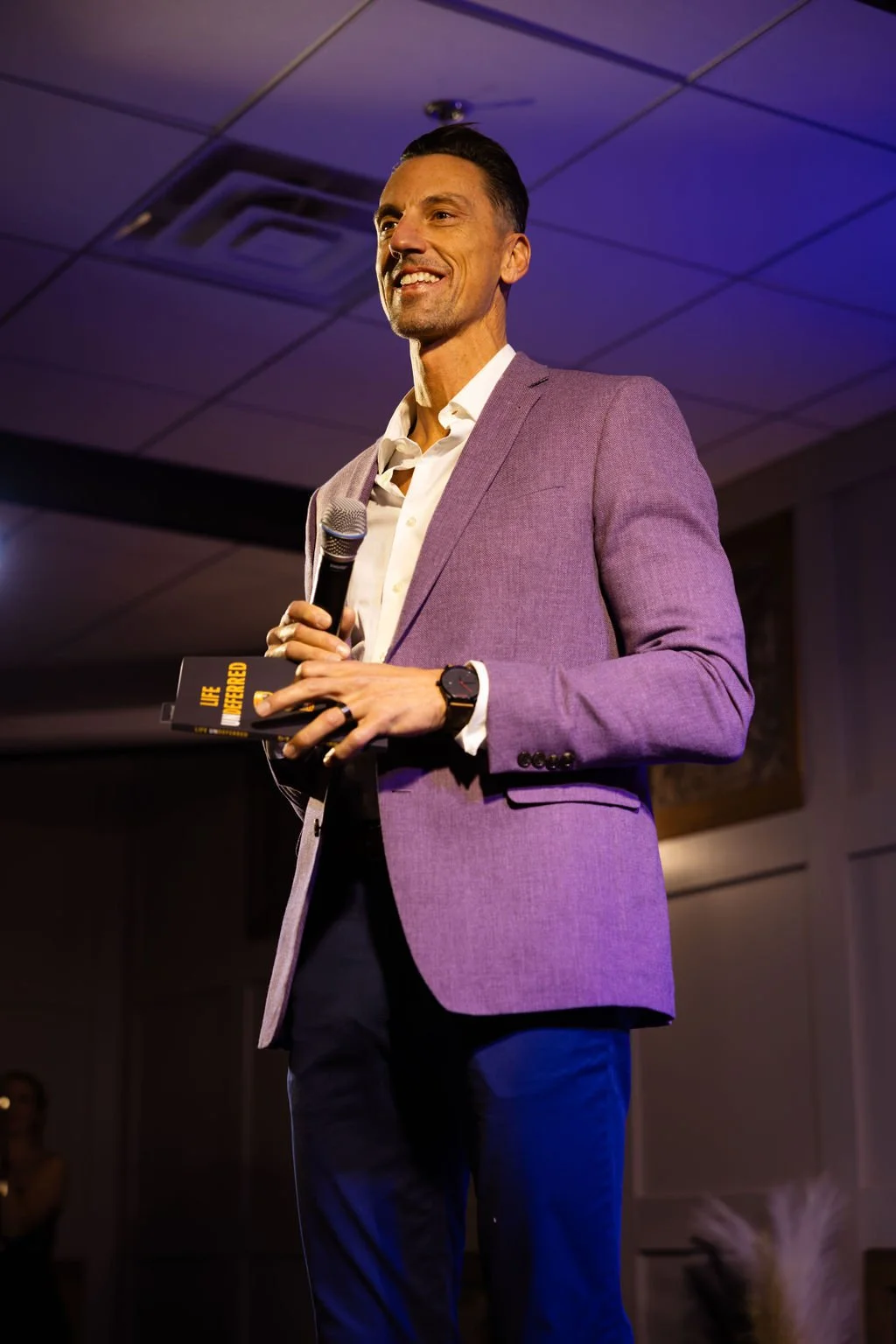 Jerrid Sebesta in a purple blazer holding a microphone and a book titled 'Life Differed' during a presentation.