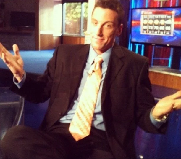 Jerrid Sebesta in a dark suit and light-colored tie sitting in a TV studio, gesturing with his hands and smiling.