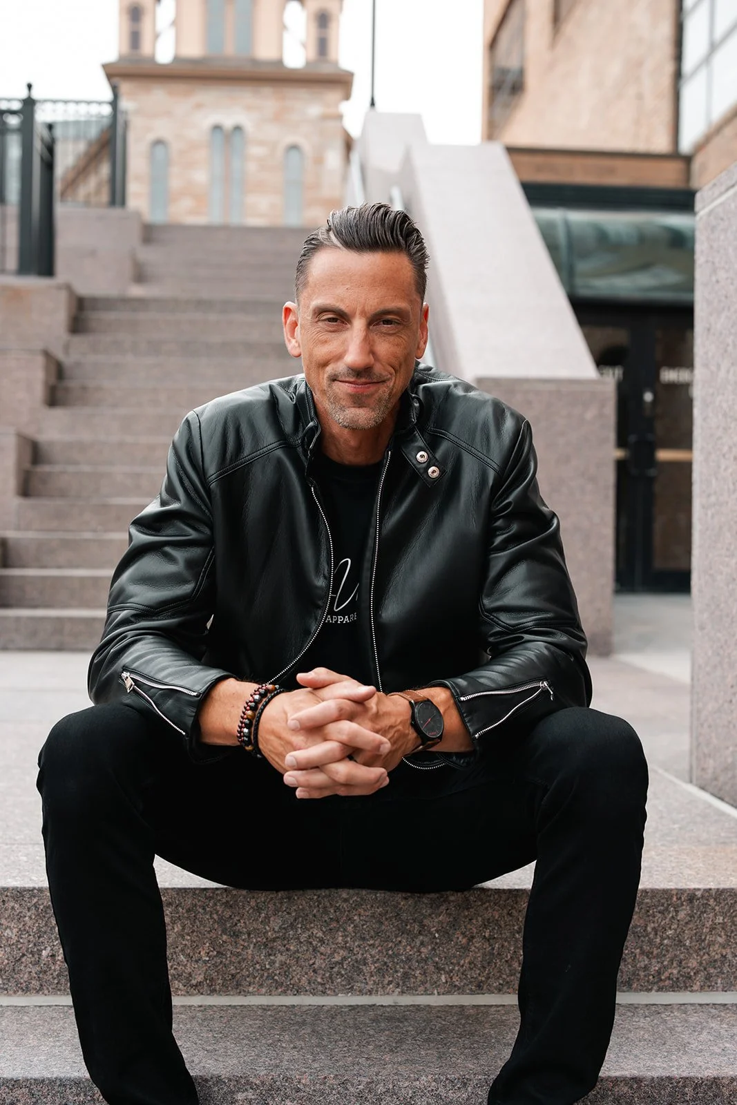 Jerrid Sebesta in a black leather jacket and black pants sitting on concrete stairs, looking at the camera with a slight smile, outdoors in an urban setting.