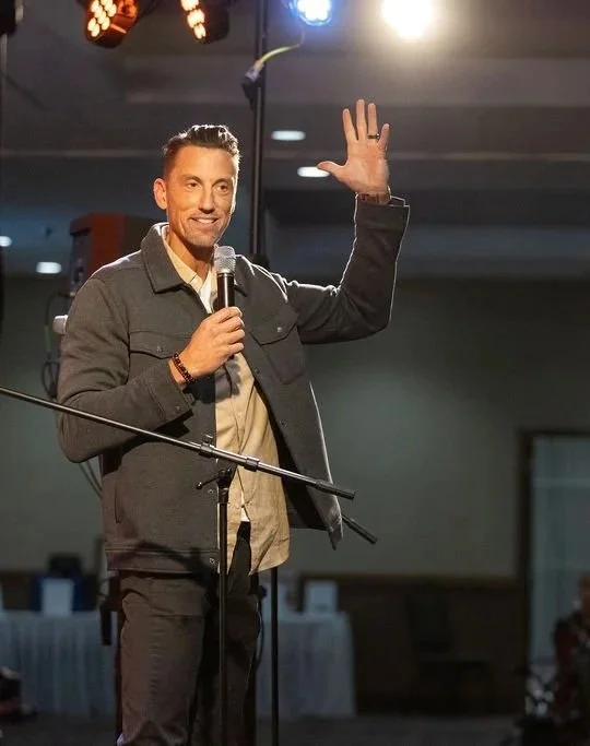 Jerrid Sebesta with short dark hair and a beard, smiling, wearing a dark jacket, holding a microphone, and waving with his right hand on stage with bright lights overhead.