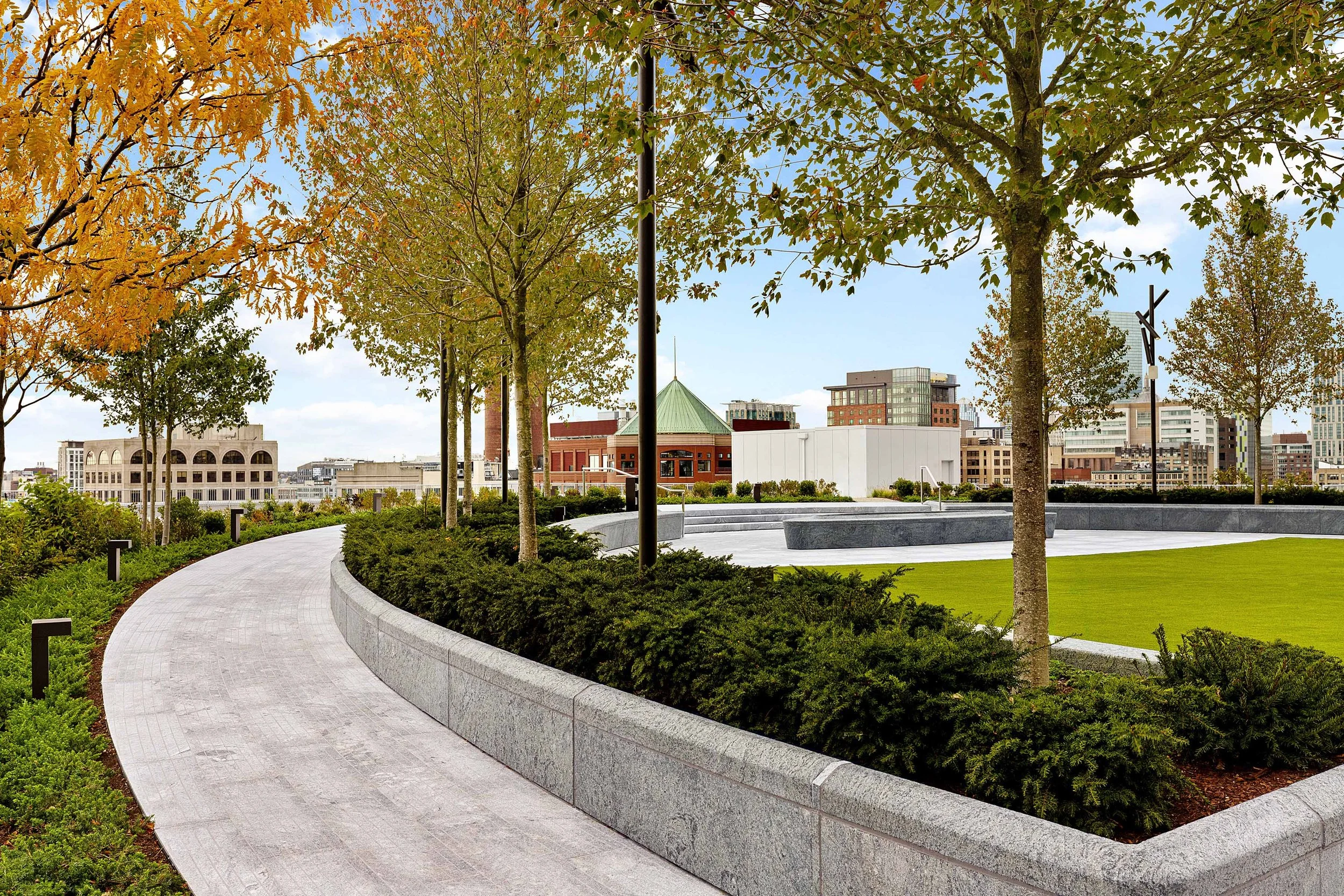 Details of the beautiful curved granite paver path at South Station Tower's Sky Park. Lush shrubbery and trees installed in vertical hardscape around the Sky Park's center oval are highlighted. In the background the oval's high performance artificial
