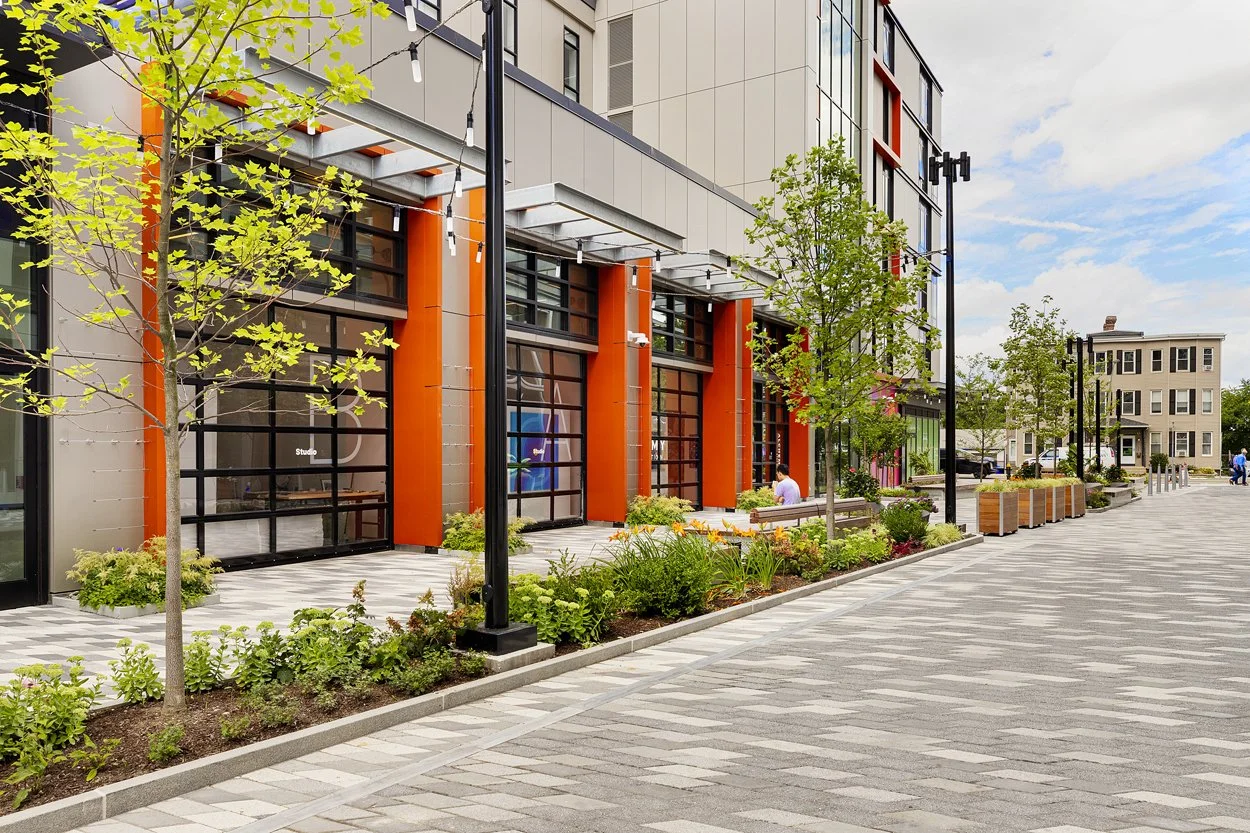 Vibrant public open space at Allston Labworks featuring modern landscape design installed by Xquisite Landscaping. The plaza includes contemporary site furnishings, wood benches, bold public art murals, new street trees, ornamental plantings, and dec