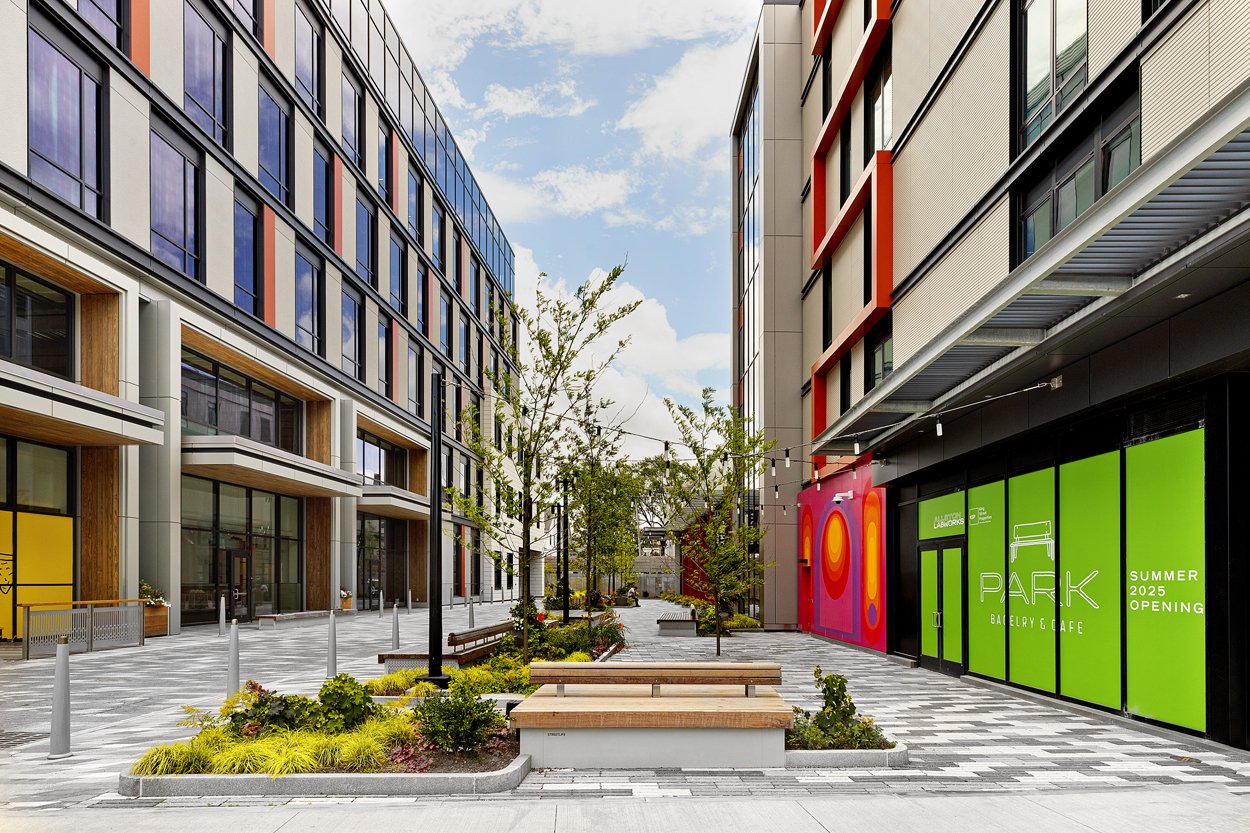 Clean, high contrast paver pathways leading into the space. Centered is a long, minimalist wooden bench integrated into a concrete planter bed filled with low-maintenance ornamental grasses and young trees.
A central, open courtyard area is visible, 