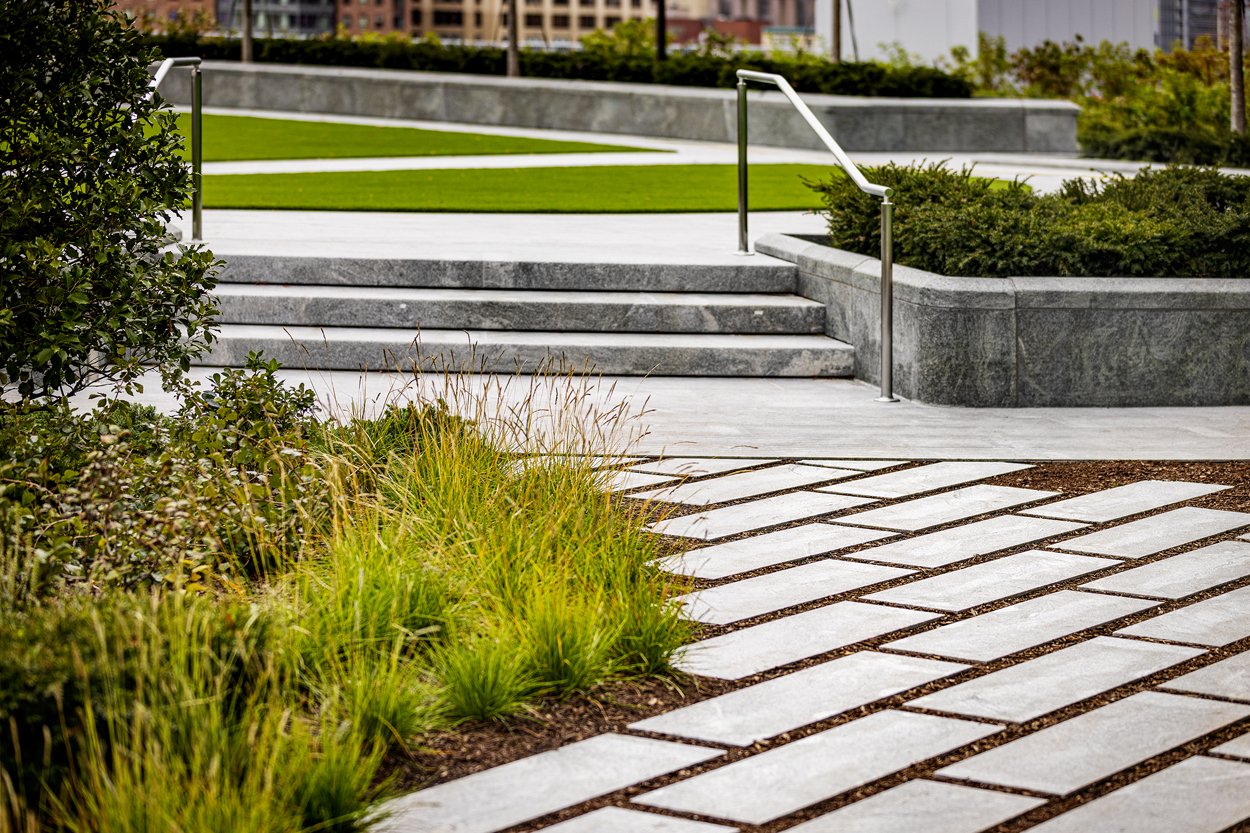 Modern rooftop park landscape at the Sky Park at South Station Tower in Boston, featuring sleek granite steps, stainless steel railings, geometric stone pavers, and lush ornamental grasses and native shrubs. The contemporary urban green space blends 