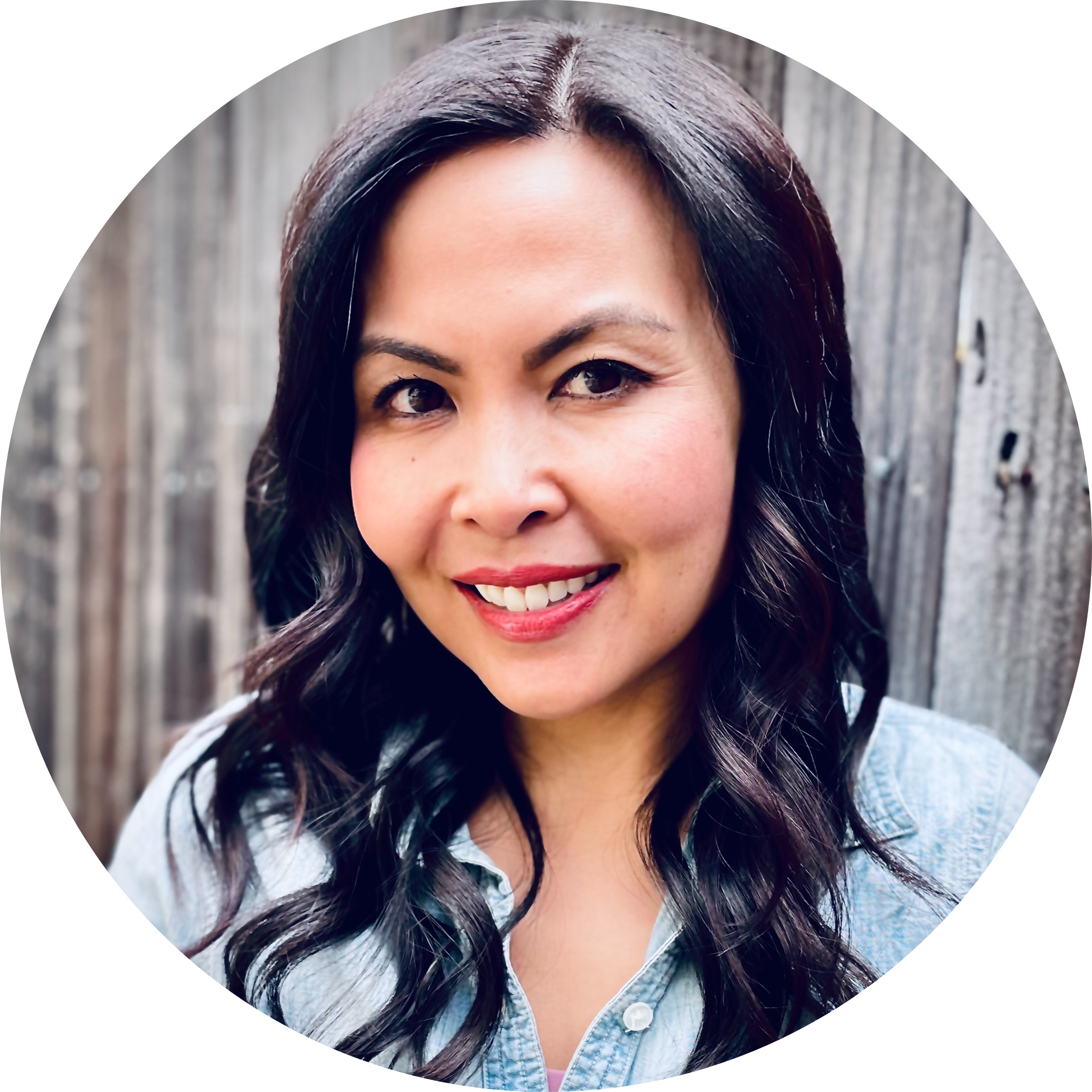 A woman with dark, wavy hair smiling outdoors in front of a wooden fence.
