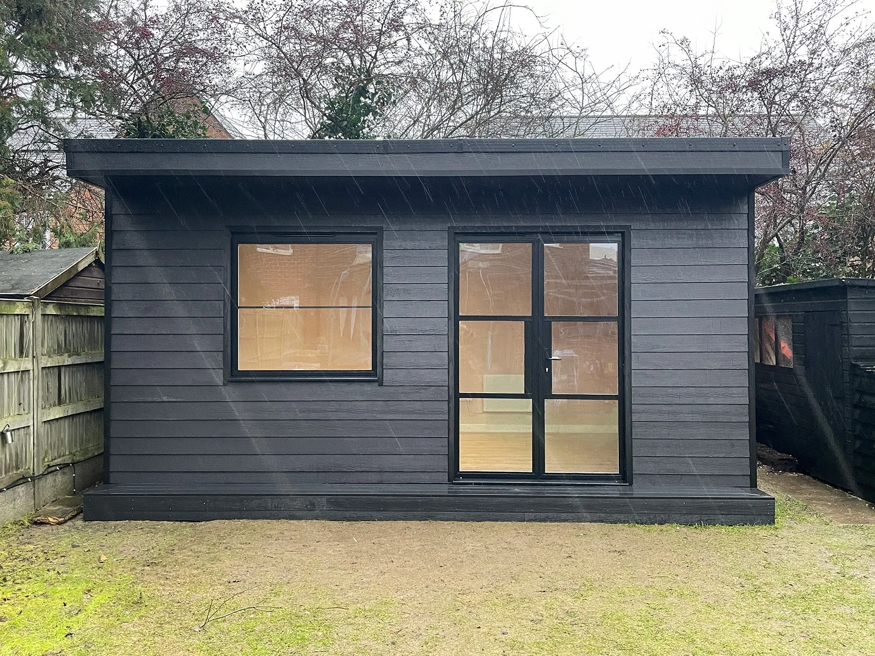 Front view of bespoke garden gym with black-framed heritage style windows and doors, black woodgrain-effect cement board cladding, a canopy and matching deck