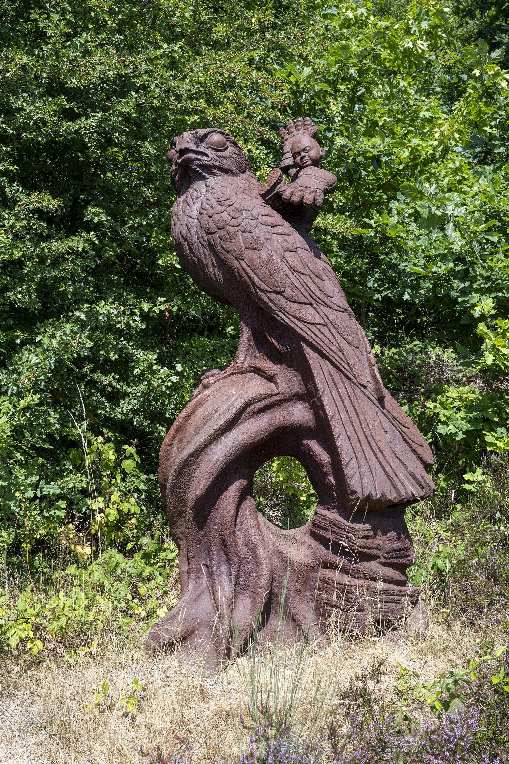 An iron sculpture of a giant kestrel with a child riding on its back wearing DIY falconer's welder's gauntlets. Perched on a twisted tree stump the bird's talons hold a claw hammer