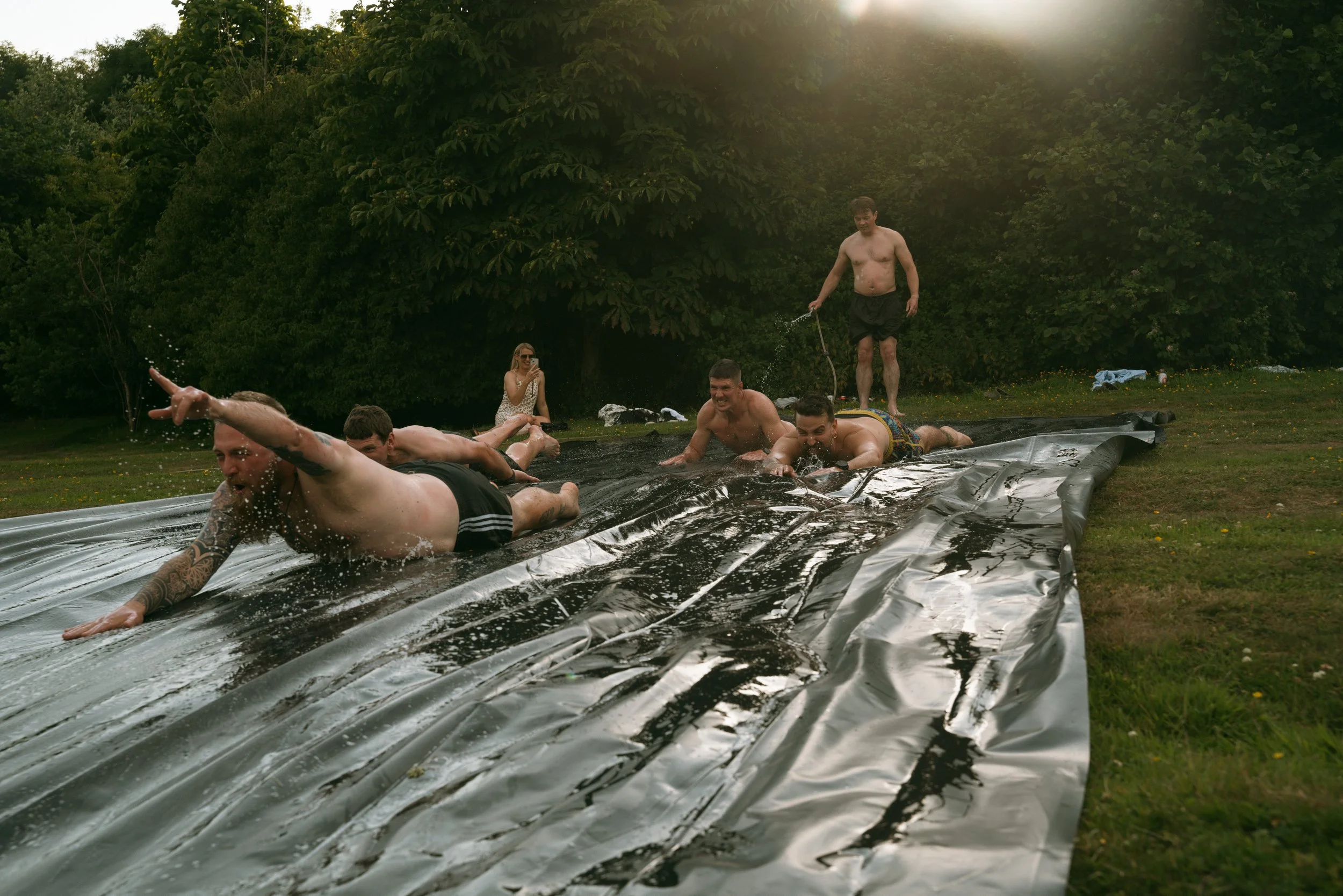 People sliding down a large water slide on a grassy field surrounded by trees during daytime.