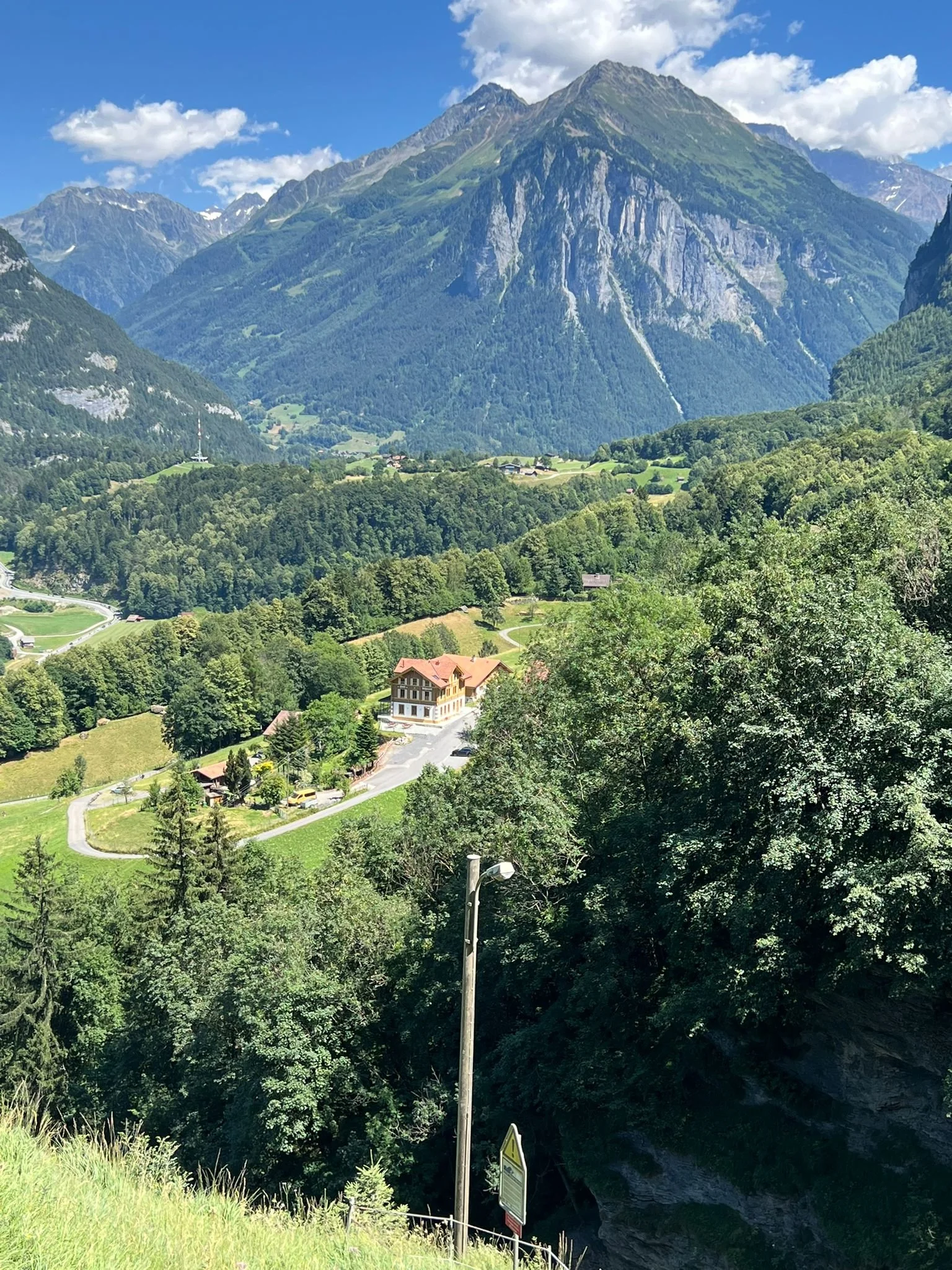 Berglandschaft mit hohen Bergen, grünen Hügeln und Wäldern, Wolken am Himmel, einige Häuser im Tal, und eine Straße mit einem Schild im Vordergrund