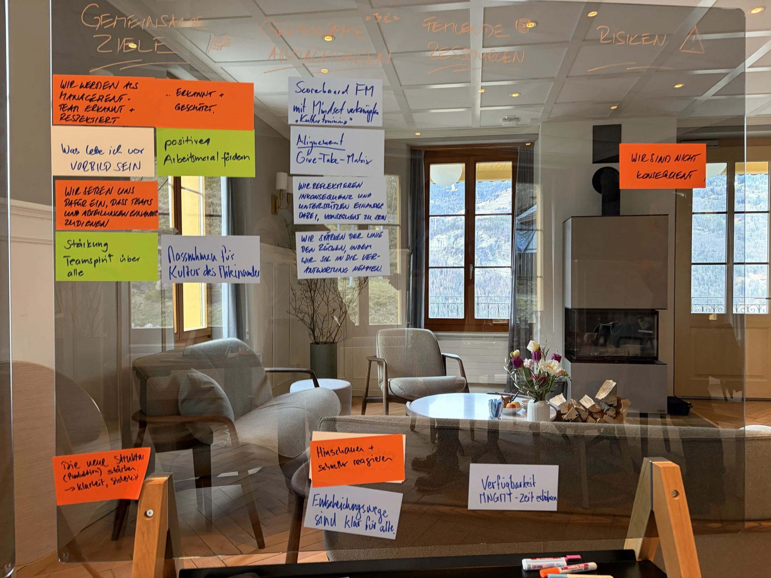 Ein Glassboard mit mehreren farbigen Zettel mit handgeschriebenen Notizen und Stichpunkten, sowie ein Wohnzimmer mit Sessel, Tisch, Blumenstrauß und Holzscheiten. Im Hintergrund sind Fenster mit Blick auf Berge und Natur.