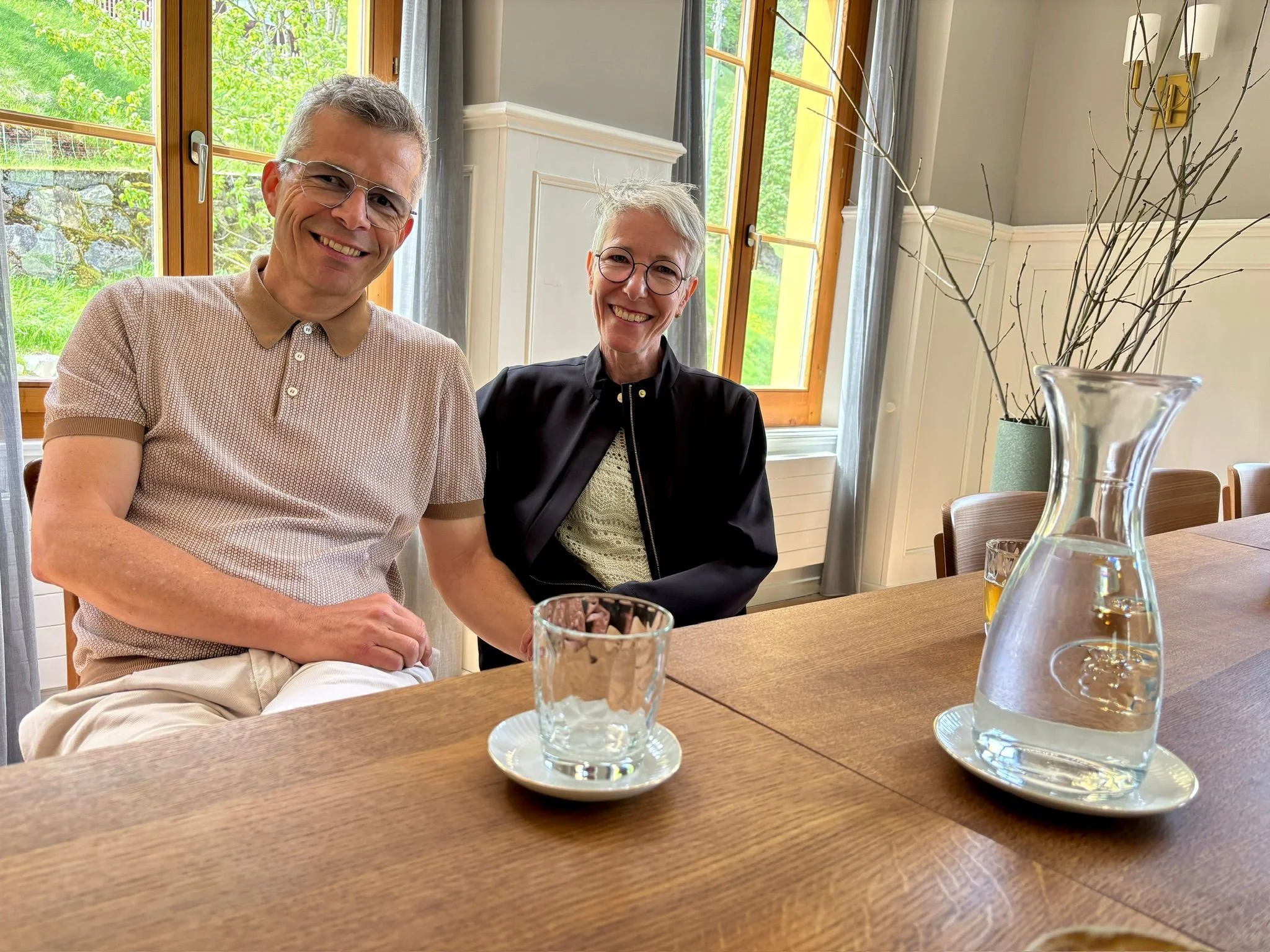 Barbara und Martin Künzi sitzen an der langen Pilgertafel in der ehemaligen Gaststube. Im Vordergrund sind ein Glas eine Karaffe Wasser auf dem Tisch zu sehen. Der Blick nach draussen zeigt grüne Bäume und einen Zaun.